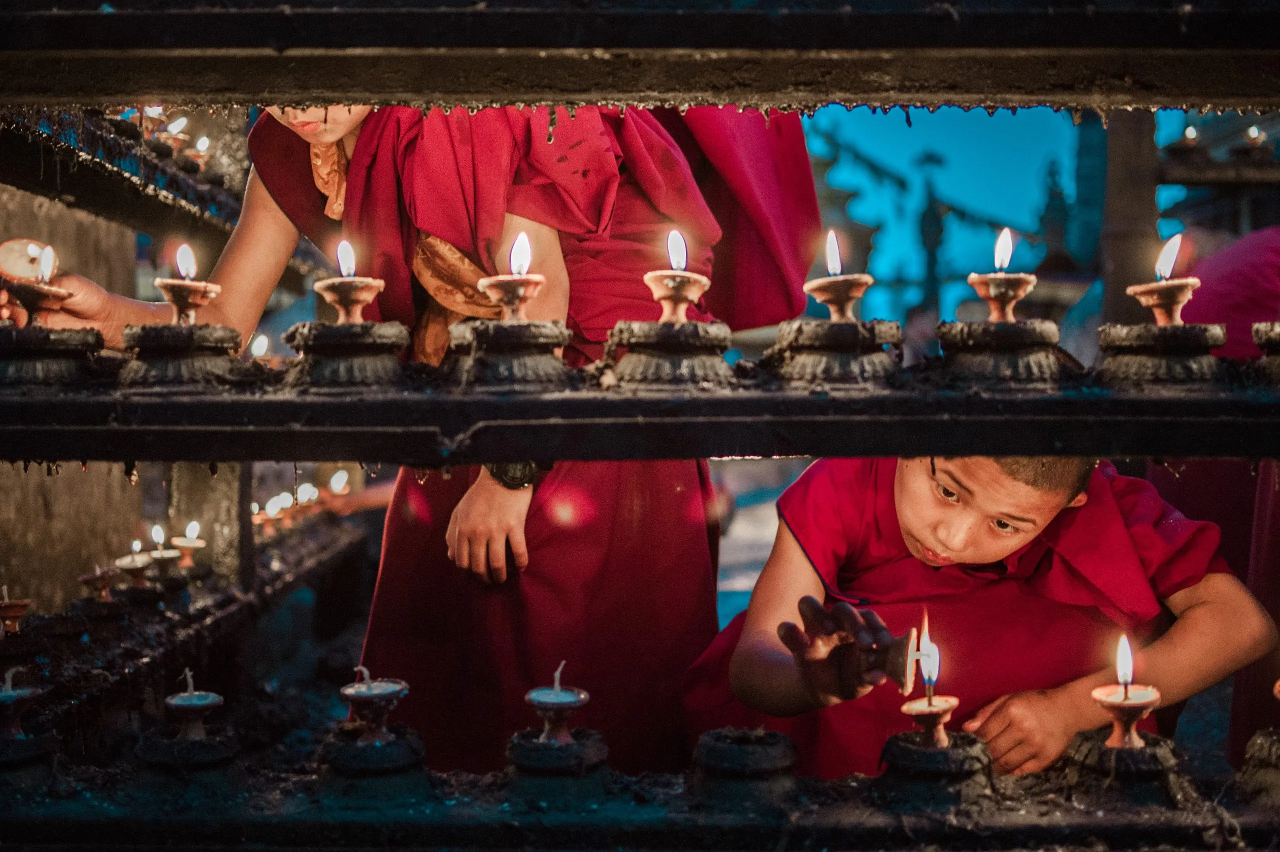 Through a black metal grill, two young monks in red robes light long rows of butter lamps at a Kathmandu monastery.