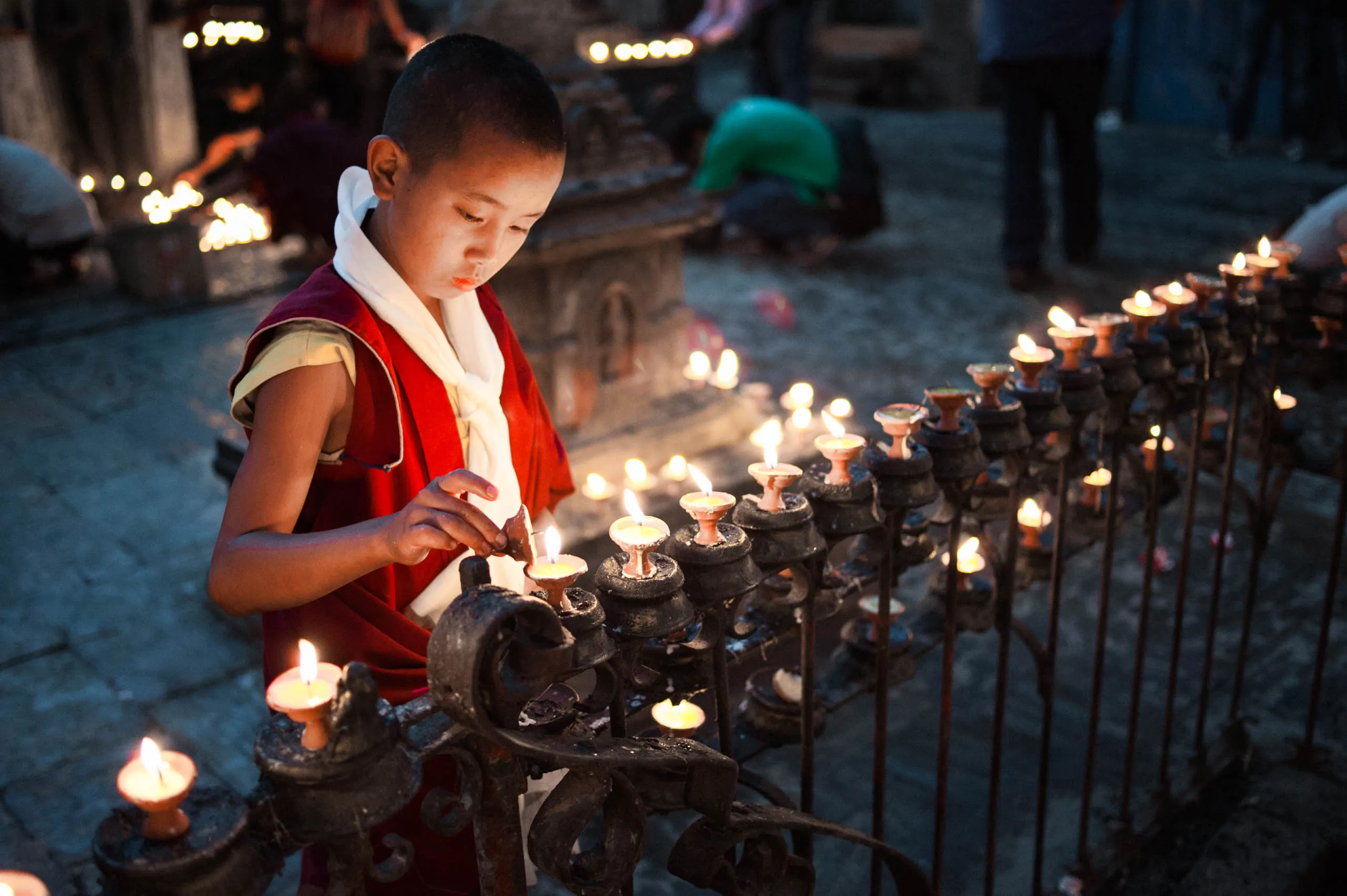 A young monk lights a row of clay butter lamps balanced along an ornate iron railing at the temple.