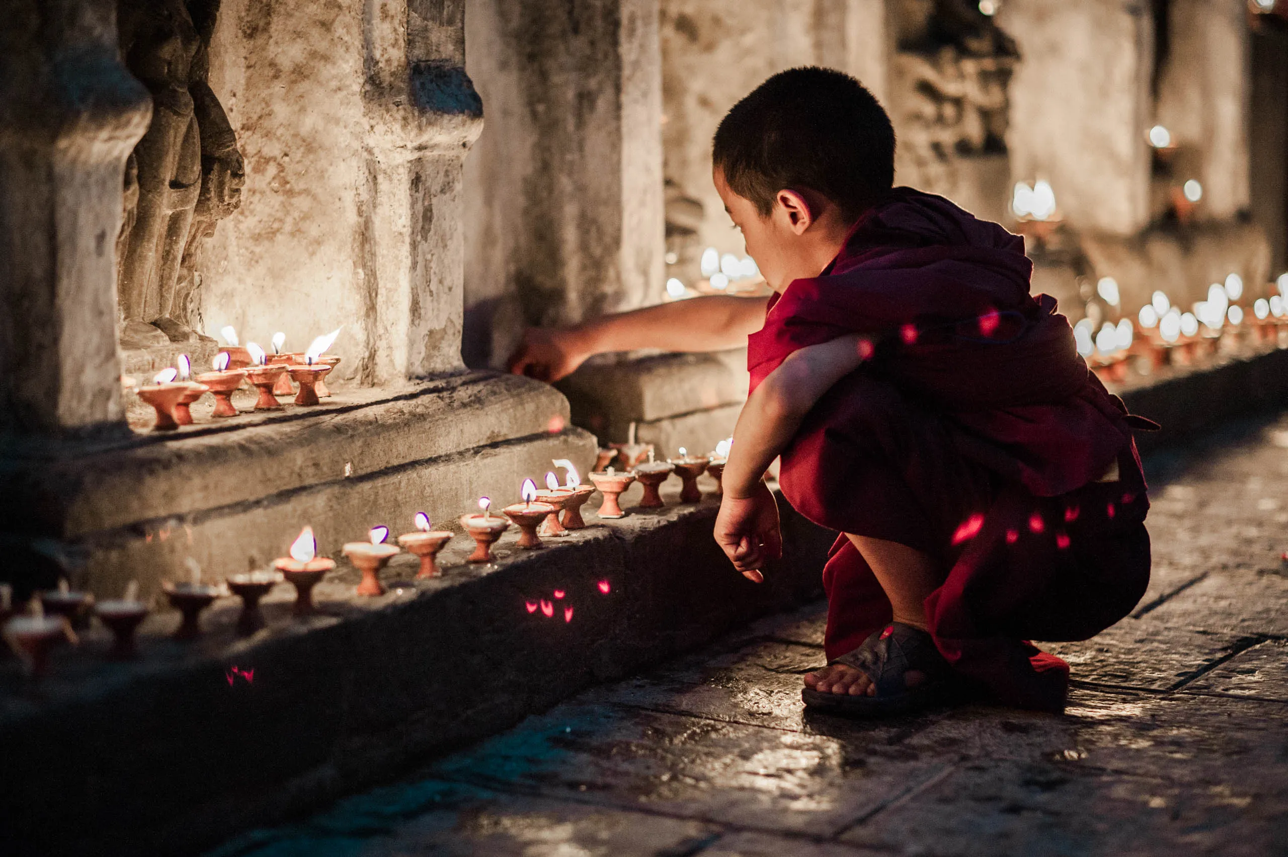A small boy monk in red lights candles at the base of a carved stone column at Swayambhunath temple.