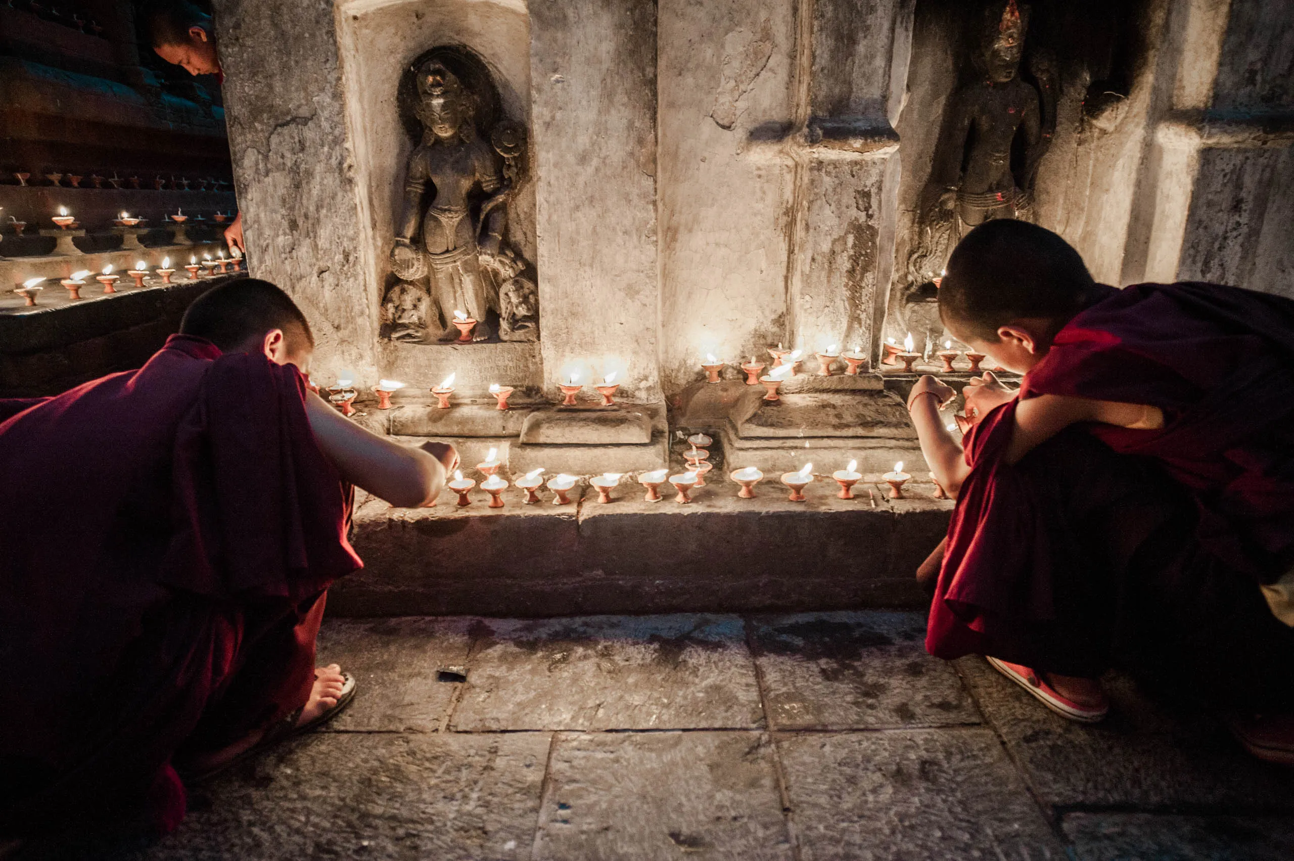 Two young monks light butter lamps in front of carved stone niches at Swayambhunath, Kathmandu.
