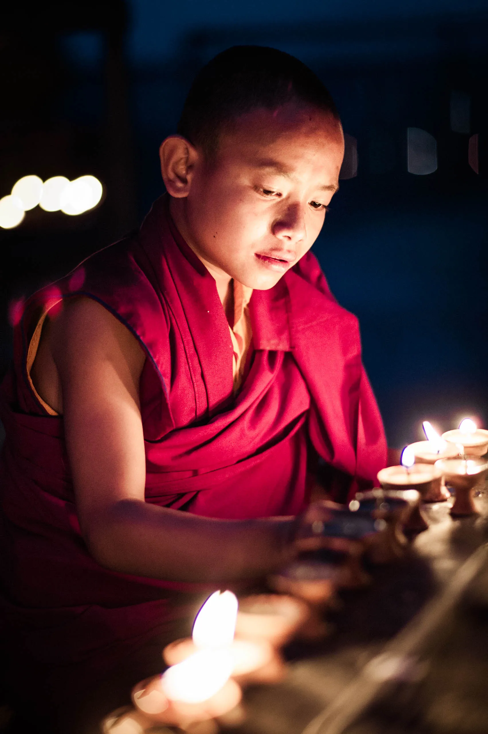 A small boy monk leans over a wooden candle stand crowded with lit butter lamps at Swayambhunath.