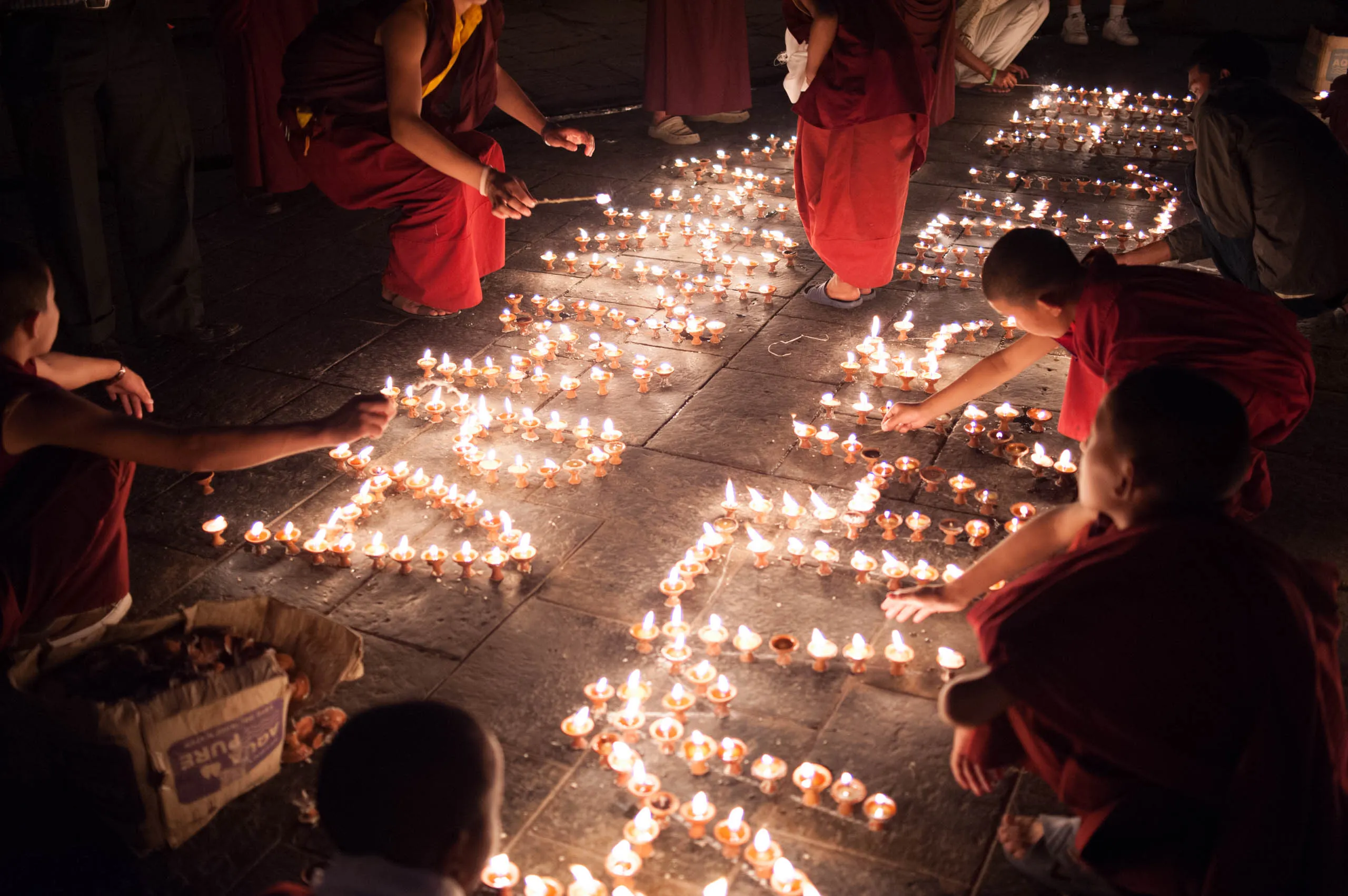 A young monk with a small backpack carefully lights a row of butter lamps along a stone wall at the temple.