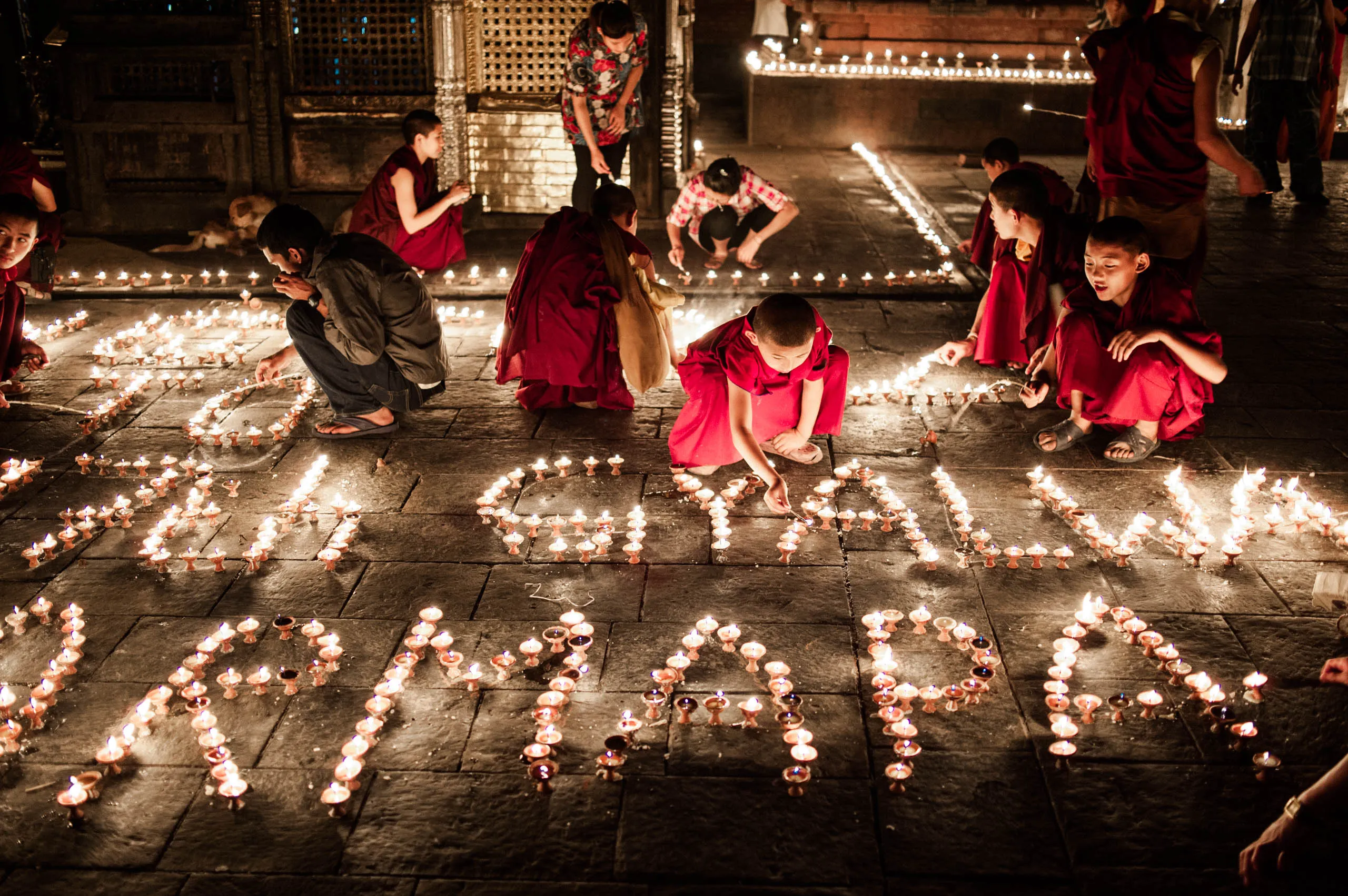 Monks of all ages bend over a long arrangement of butter lamps shaped into letters across the temple courtyard.