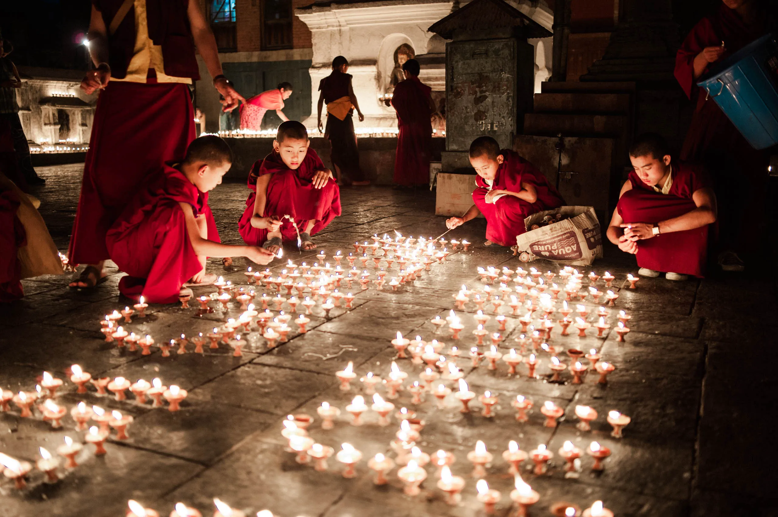 A close portrait of a young monk in red robes, with butter lamps glowing in the foreground at twilight.