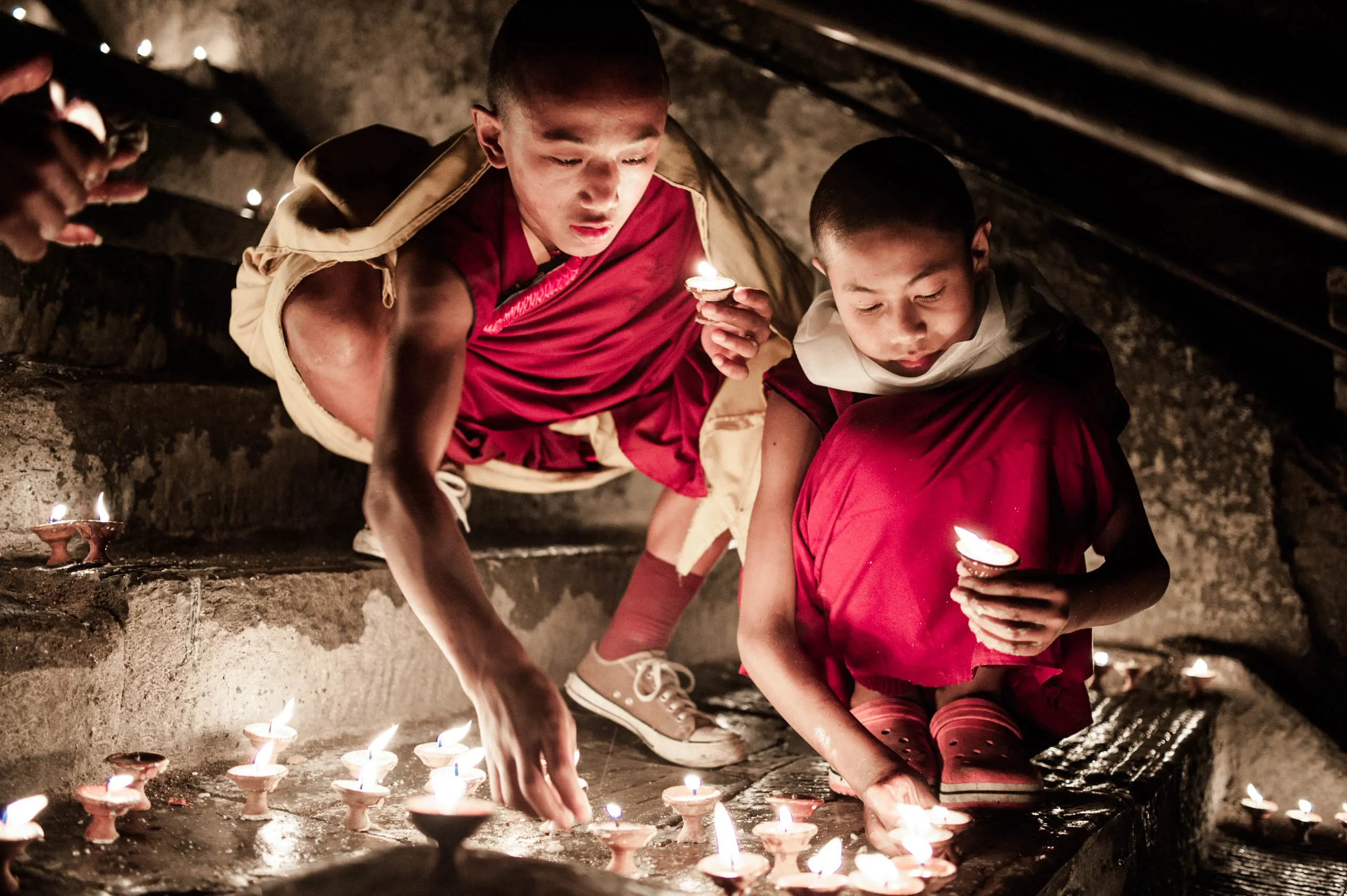 Two boy monks lean forward to light candles together inside the temple at Swayambhunath, Kathmandu.