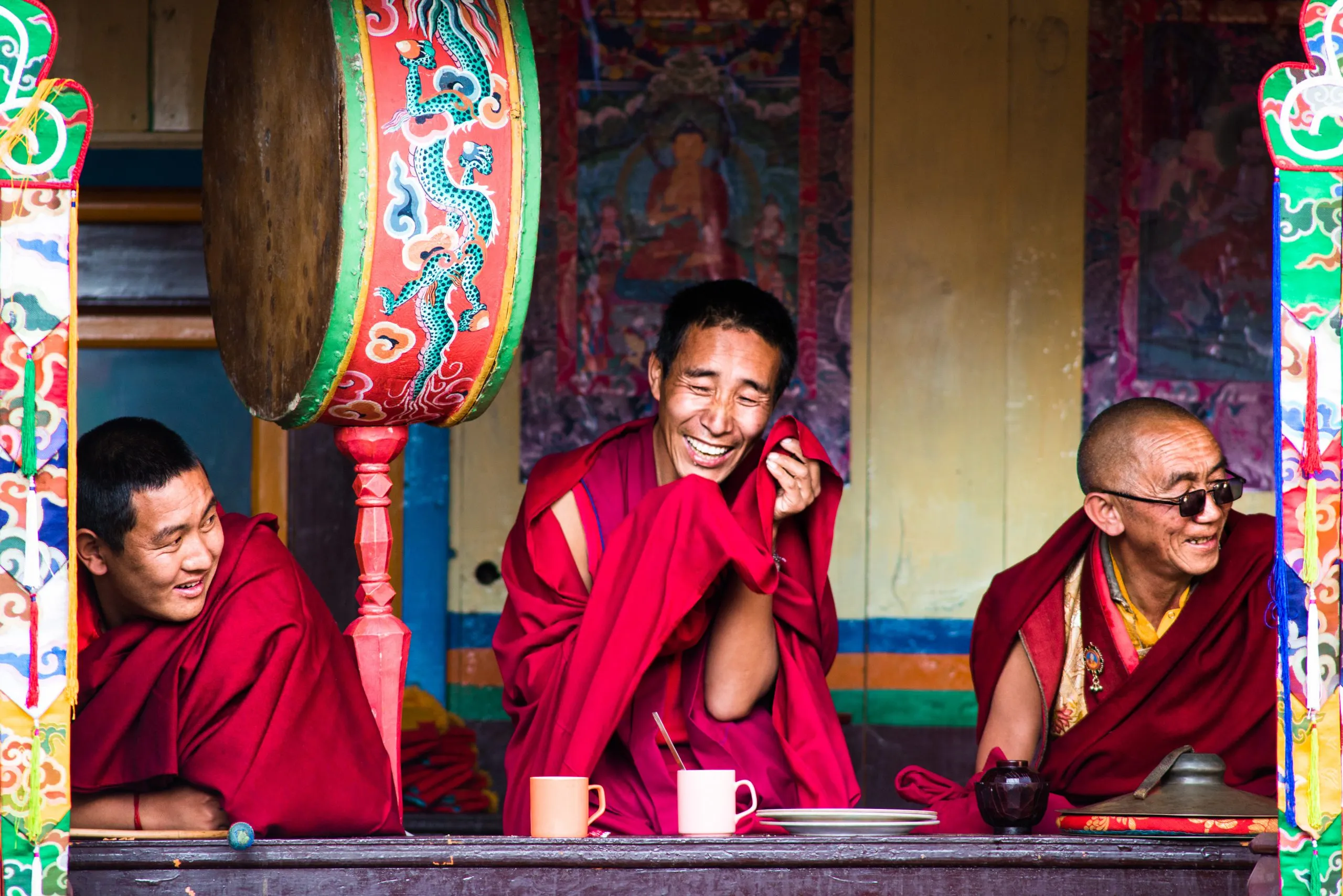 Three Tibetan Buddhist monks in maroon robes laugh together at the window of a Nepali monastery beside a painted ceremonial drum.