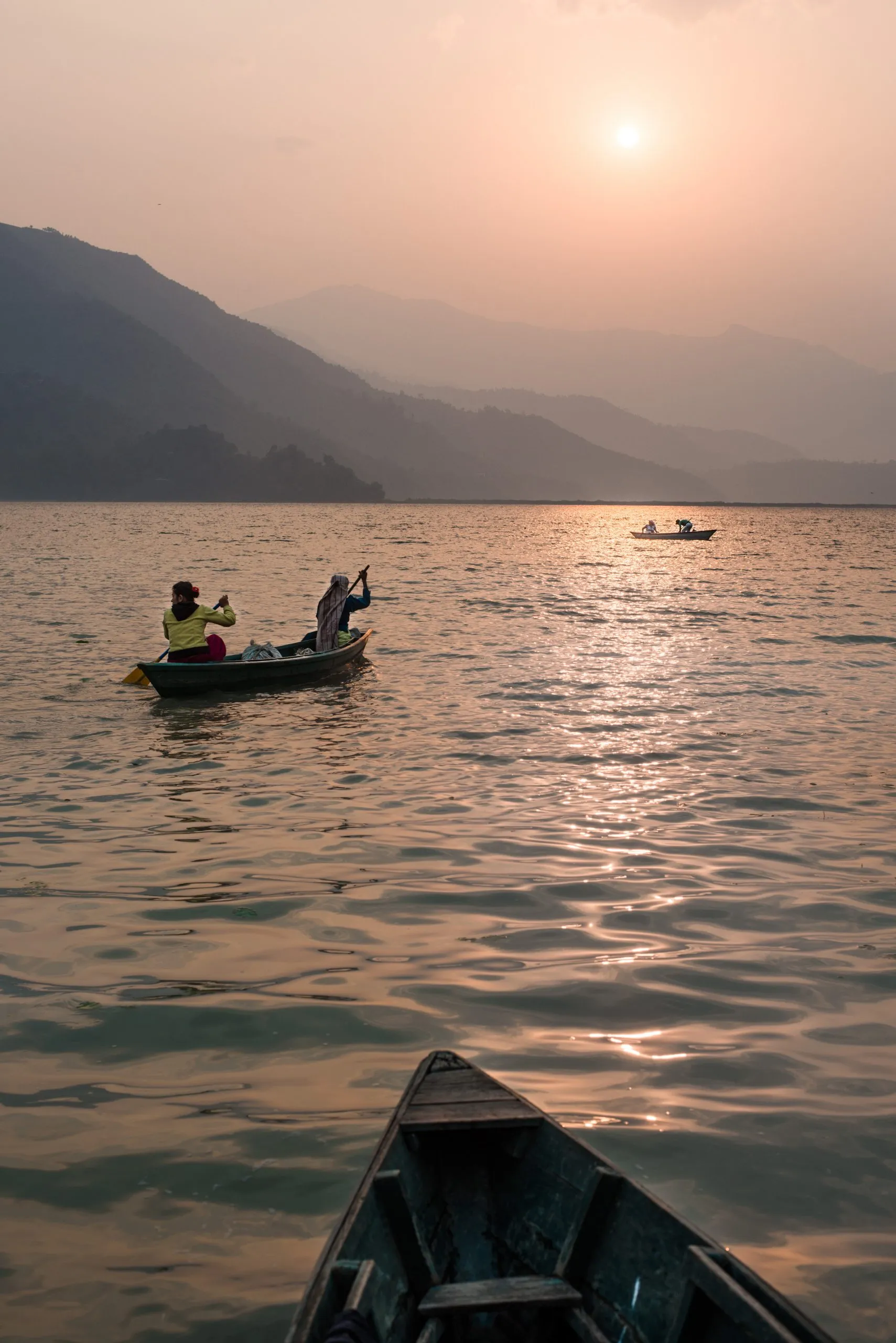 Two people row a wooden boat on Phewa Lake at sunset in Pokhara, Nepal, with hazy hills layered behind.