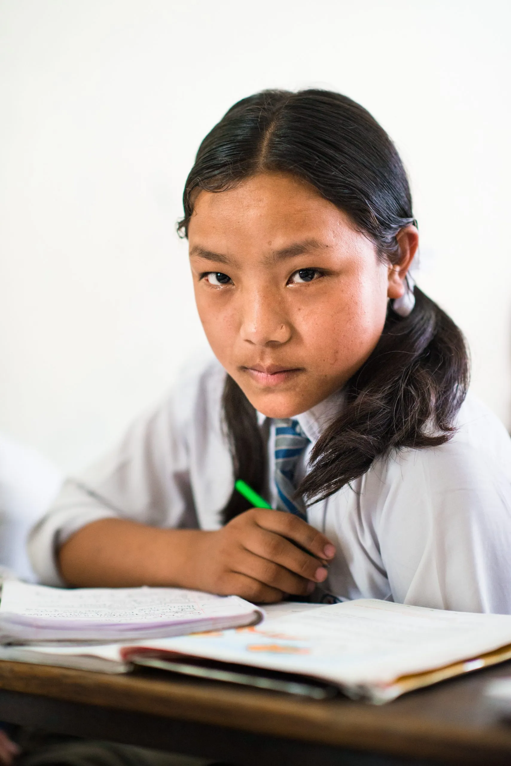 Portrait of a Nepali schoolgirl in a white shirt and tie, looking at the camera with an open notebook and a green pen in her hand.