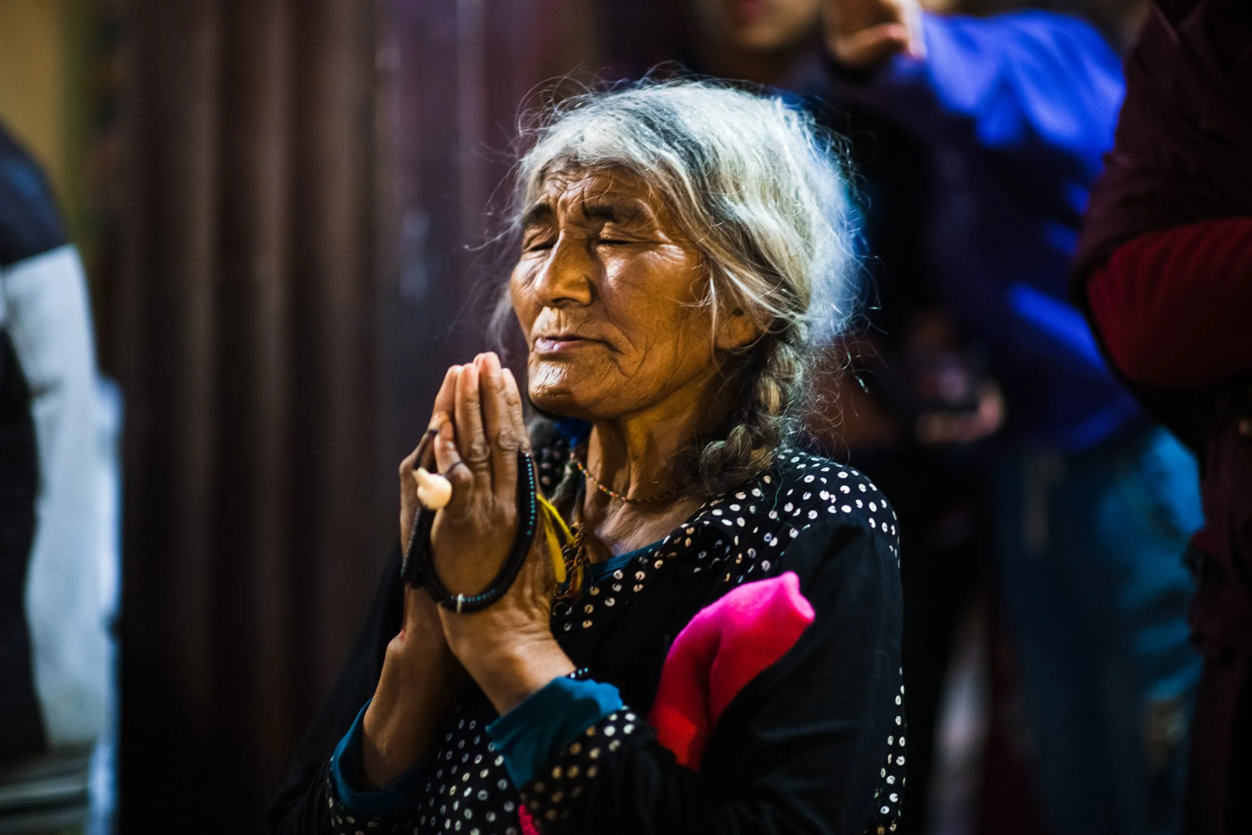 An older Nepali woman with grey hair and prayer beads pressed her palms together in prayer, eyes closed, a small votive between her hands.