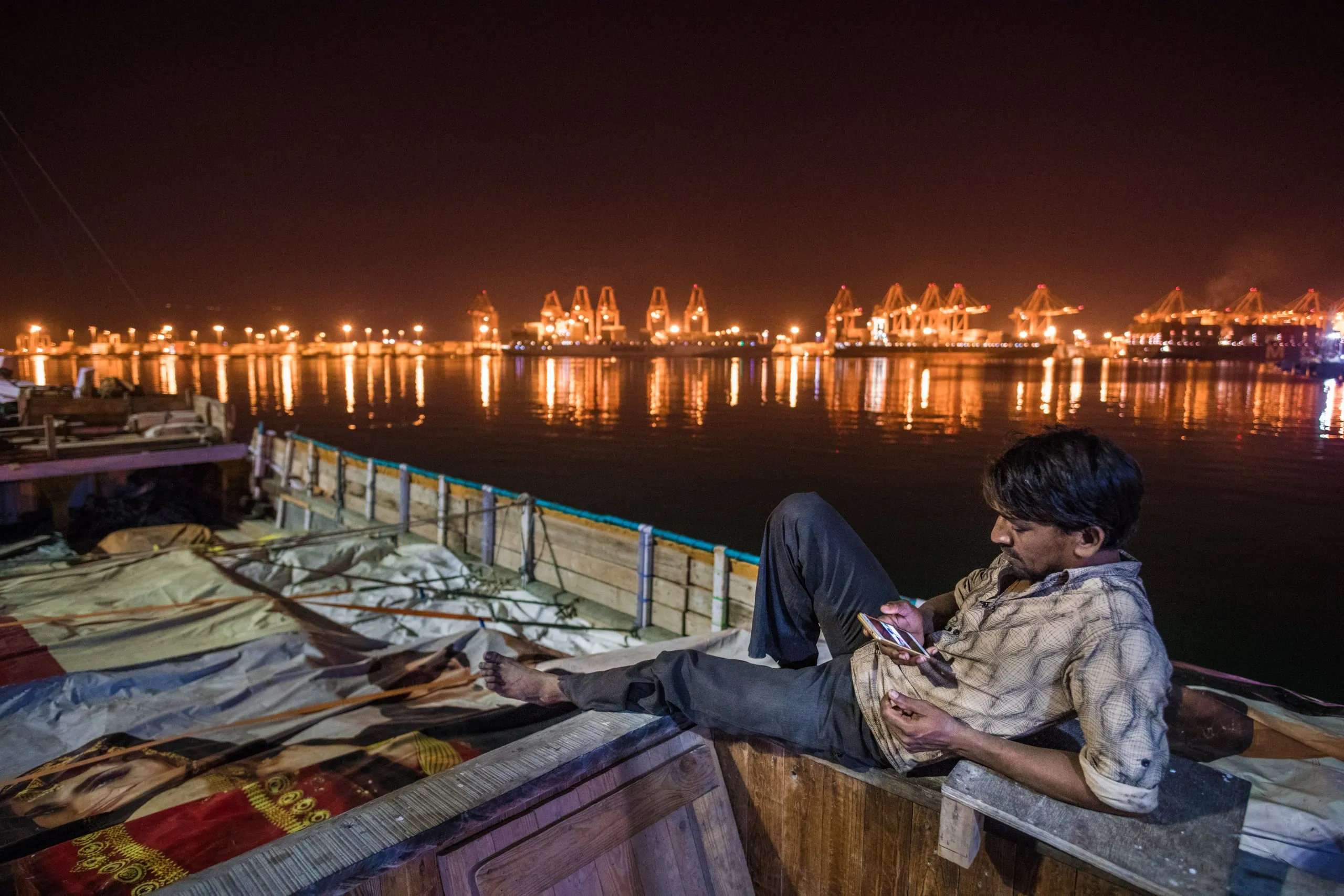 A man reclines on the deck of a wooden cargo dhow at night in Salalah port, Oman, with the lit cranes of the container terminal reflected in the water behind.