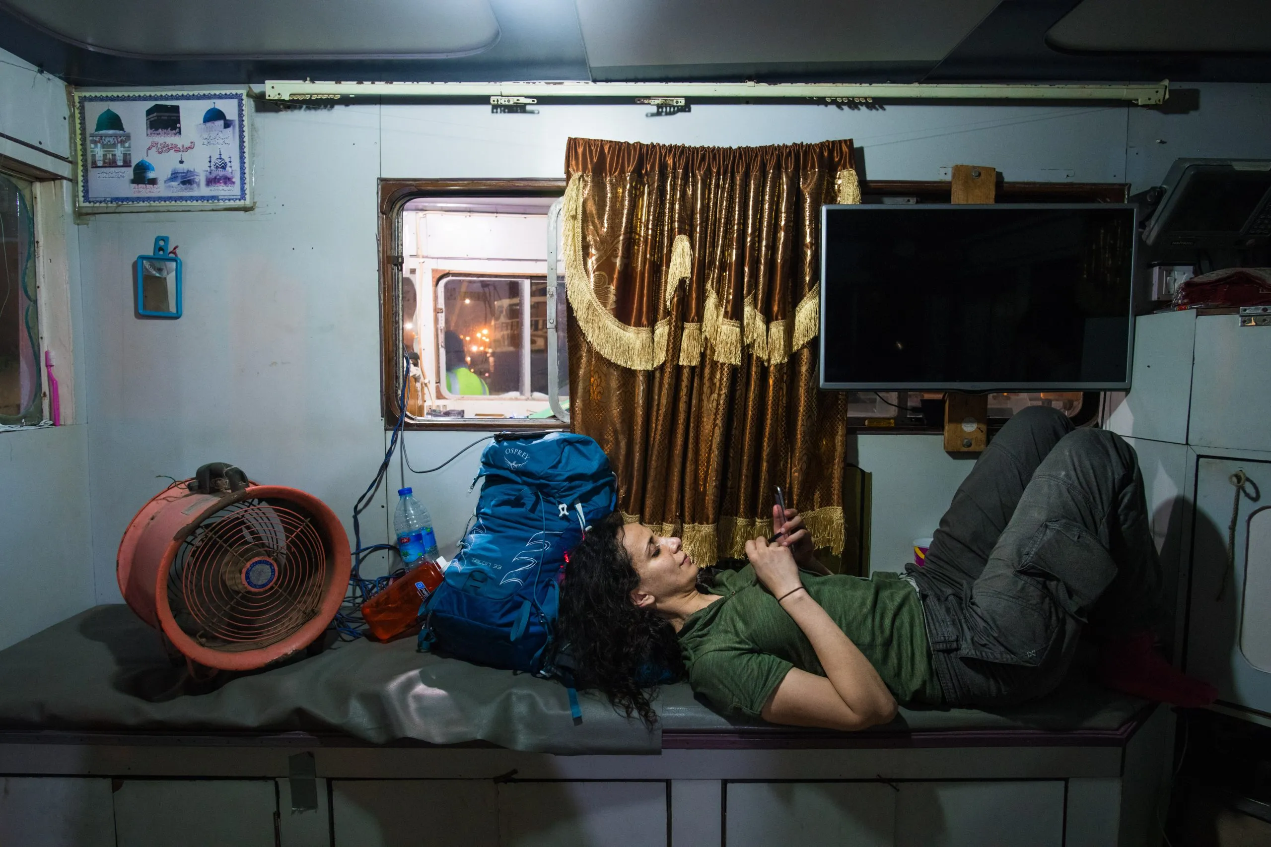 A traveler lies on a bench inside the cabin of a wooden cargo dhow about to depart from Oman for Socotra, blue backpack and orange fan beside her.