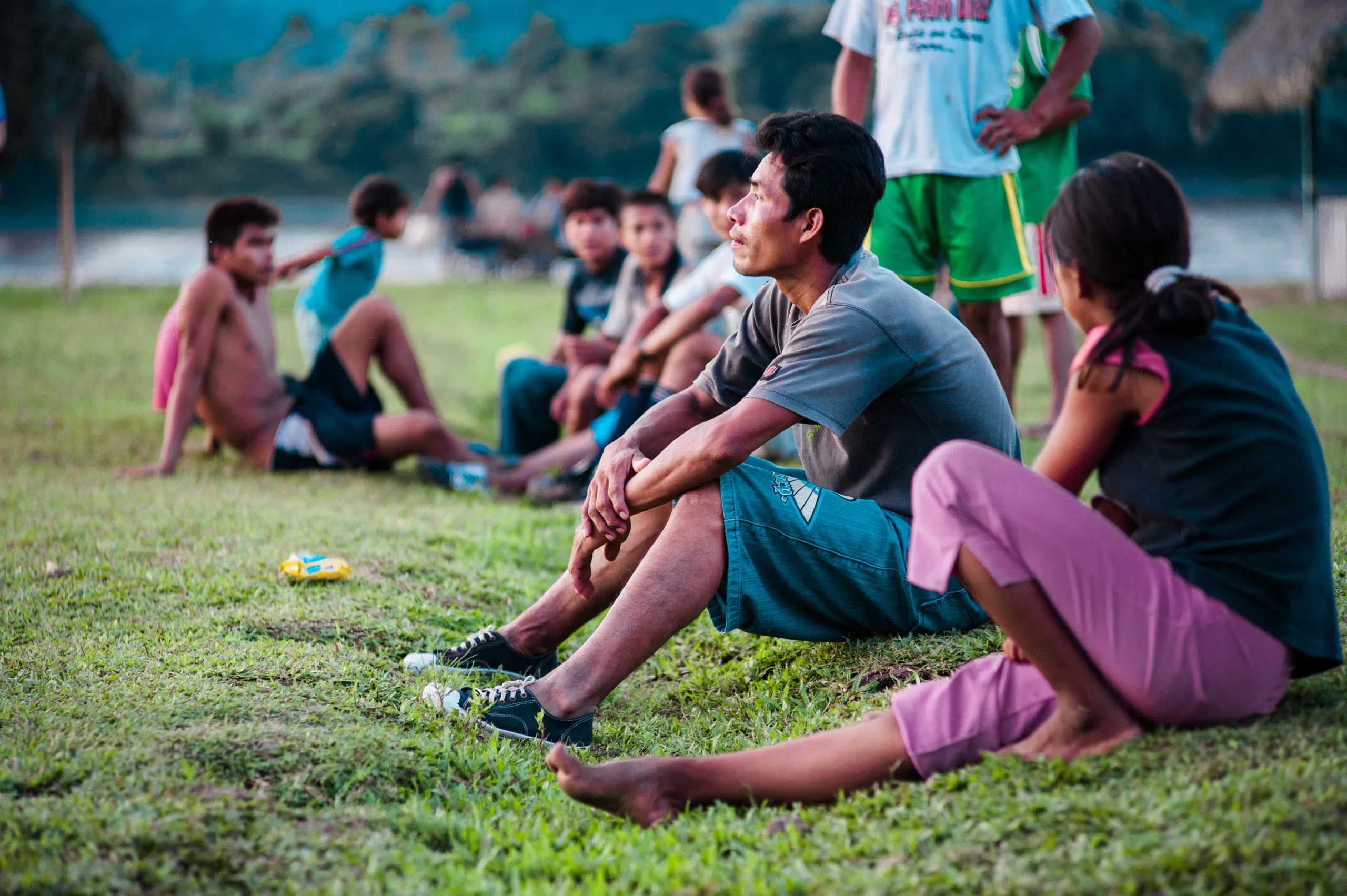 A group of Indigenous Amazonian community members sit on the grass watching a football match at dusk in eastern Peru.