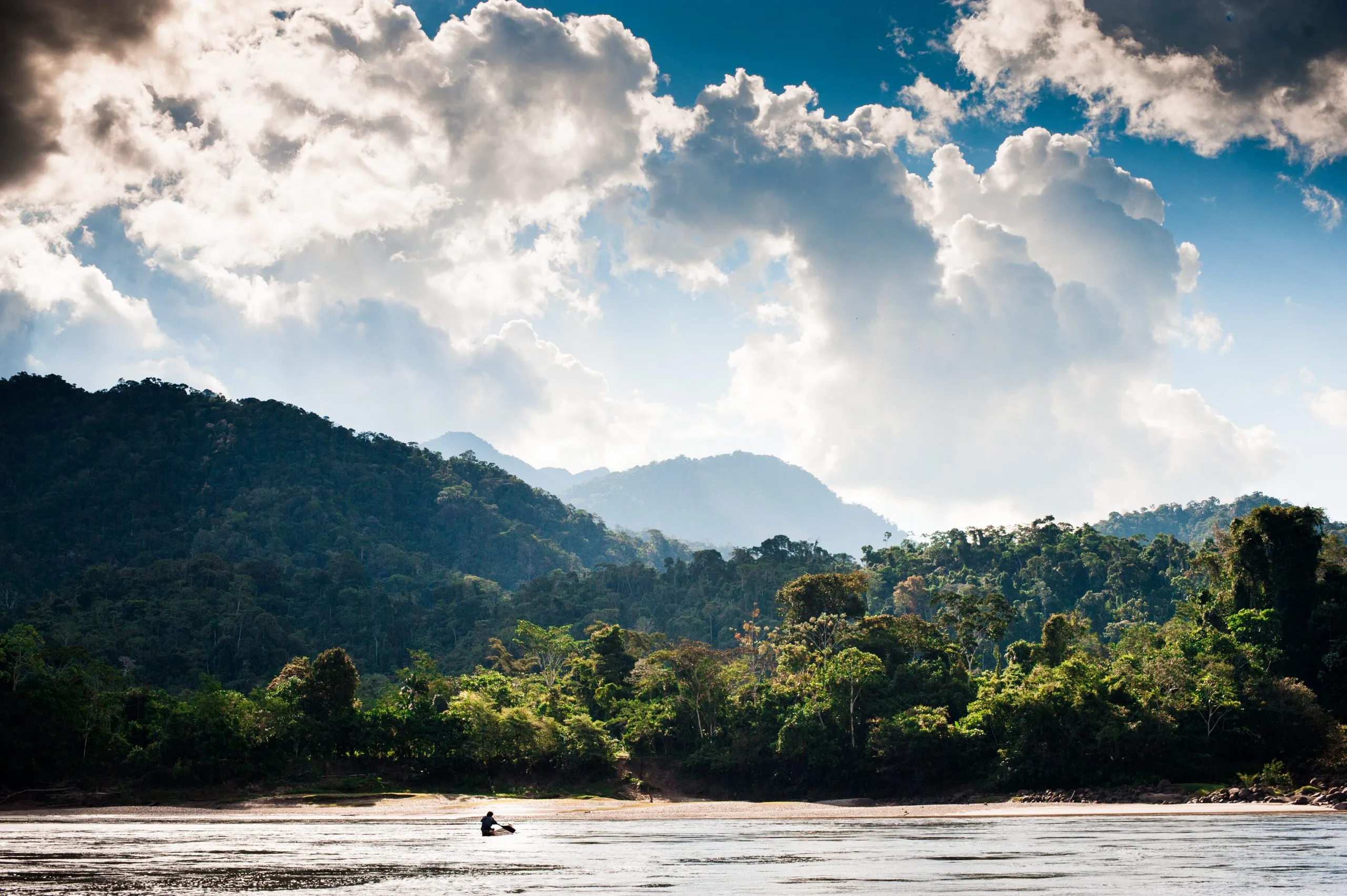 A small boat moves down a wide brown Amazon tributary in Peru, dense rainforest mountains rising under towering tropical clouds.