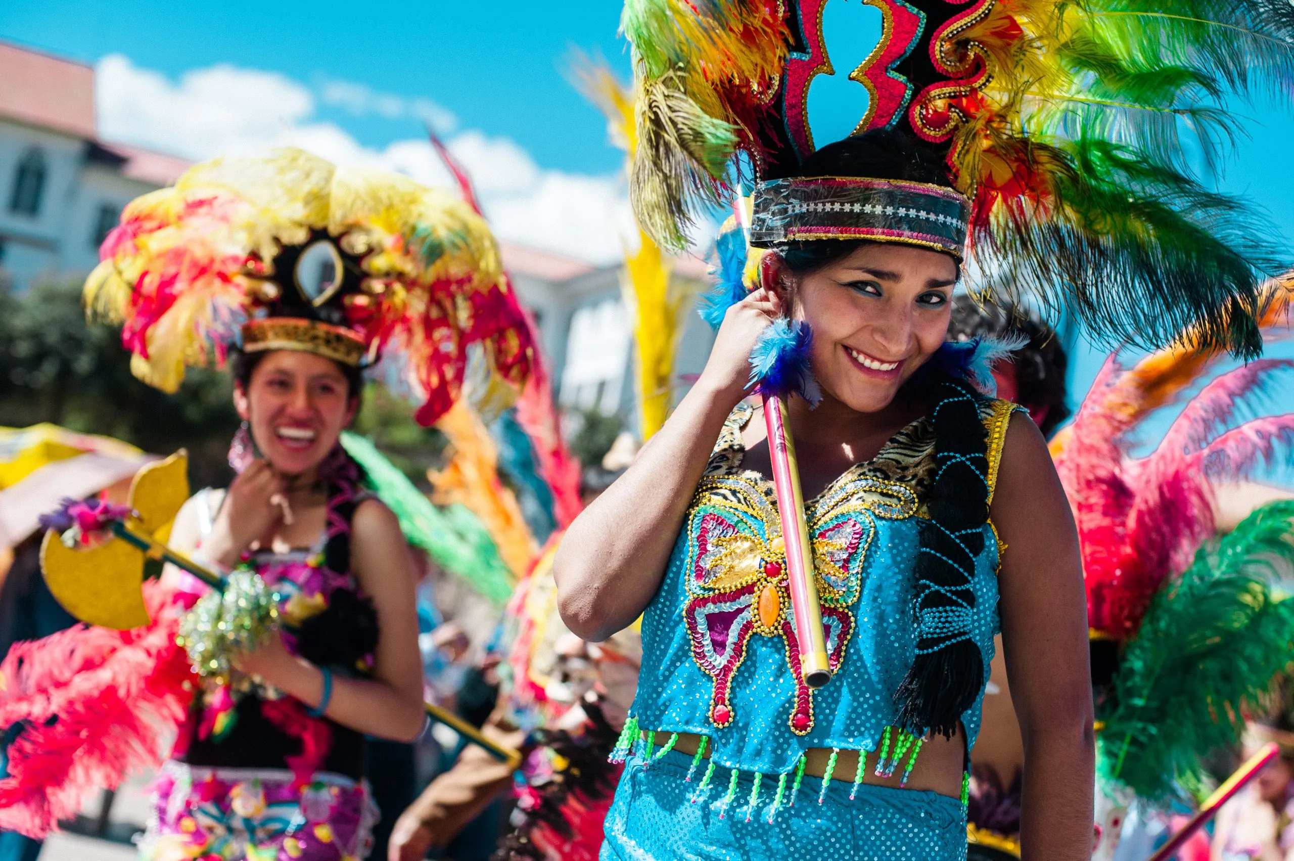 Two women in vibrant feathered carnival headdresses and beaded costumes smile during a traditional festival parade in Peru.