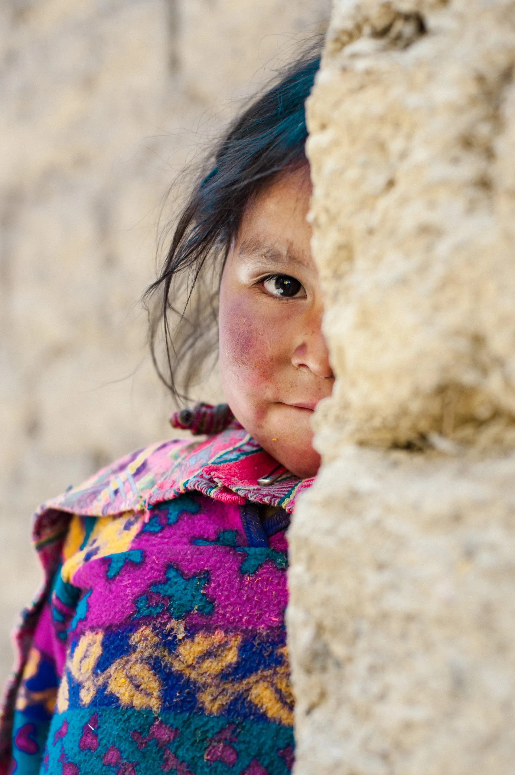 An Andean girl in a vivid pink-and-blue patterned poncho peeks out from behind an adobe wall, looking shyly at the camera.