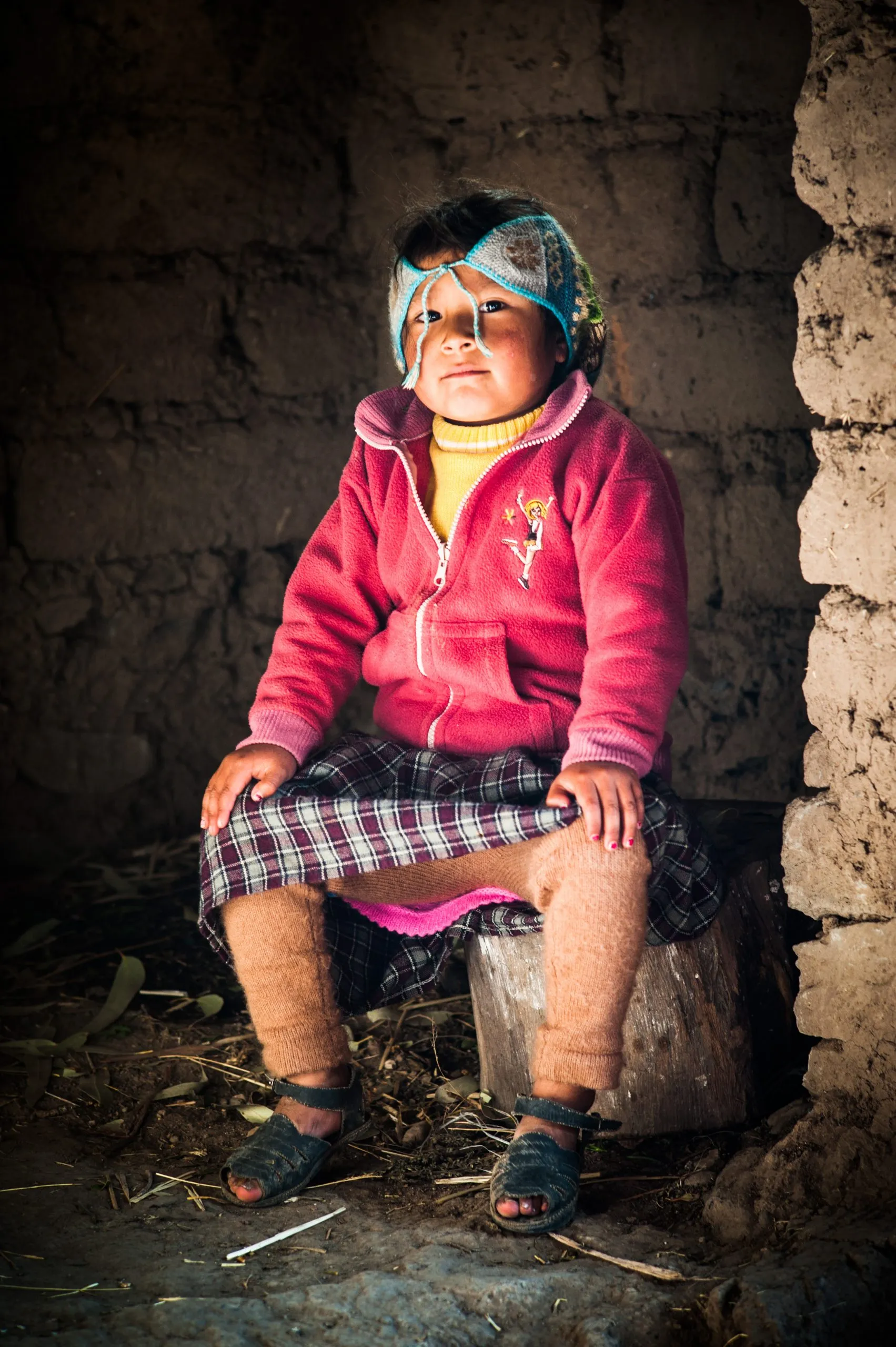 Portrait of an Andean child in a colourful patterned poncho and woven cap, sitting on a stool against an adobe wall in the Peruvian highlands.
