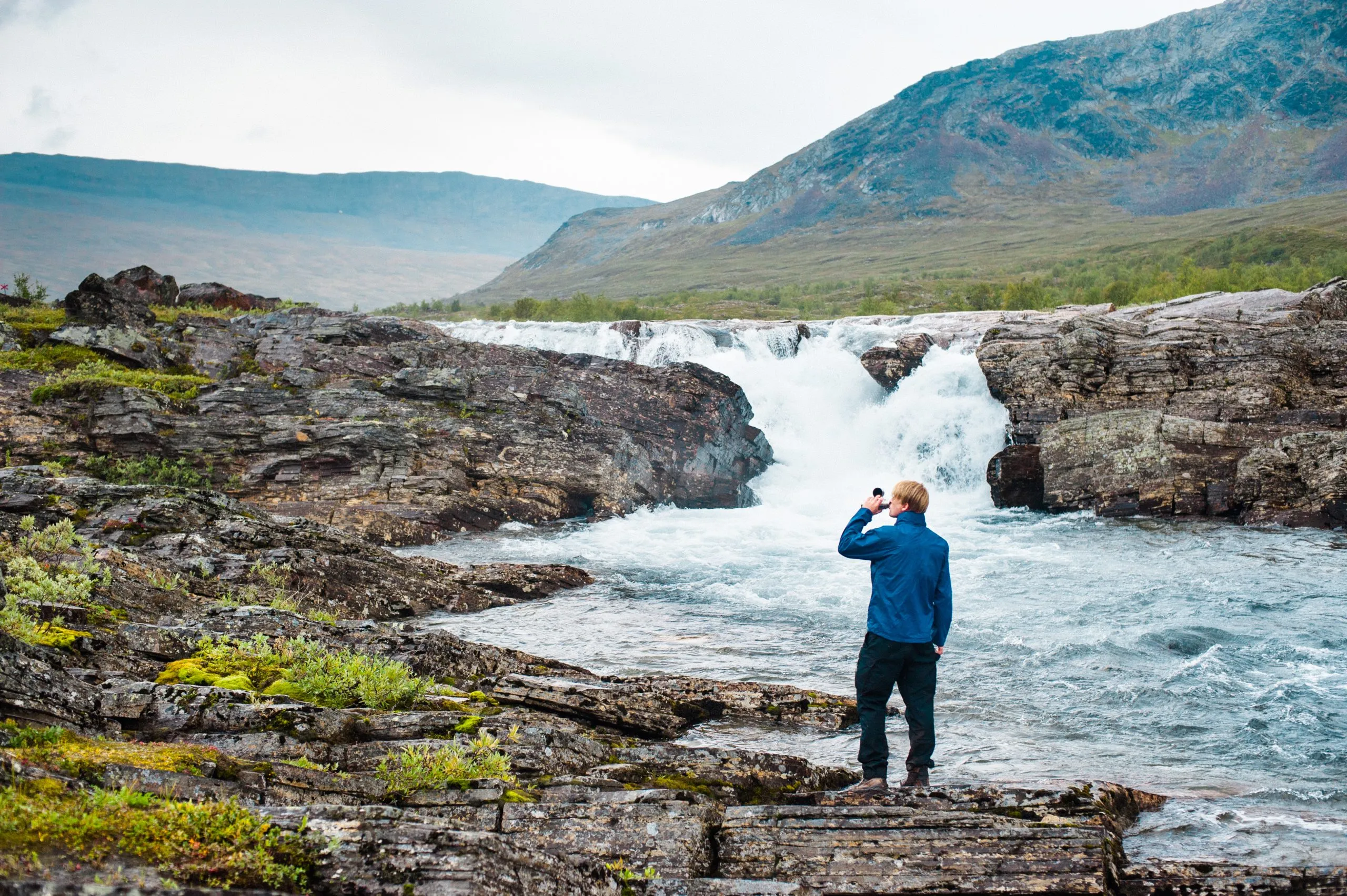 A person in a blue jacket photographs a roaring river waterfall in the Swedish Lapland mountains.