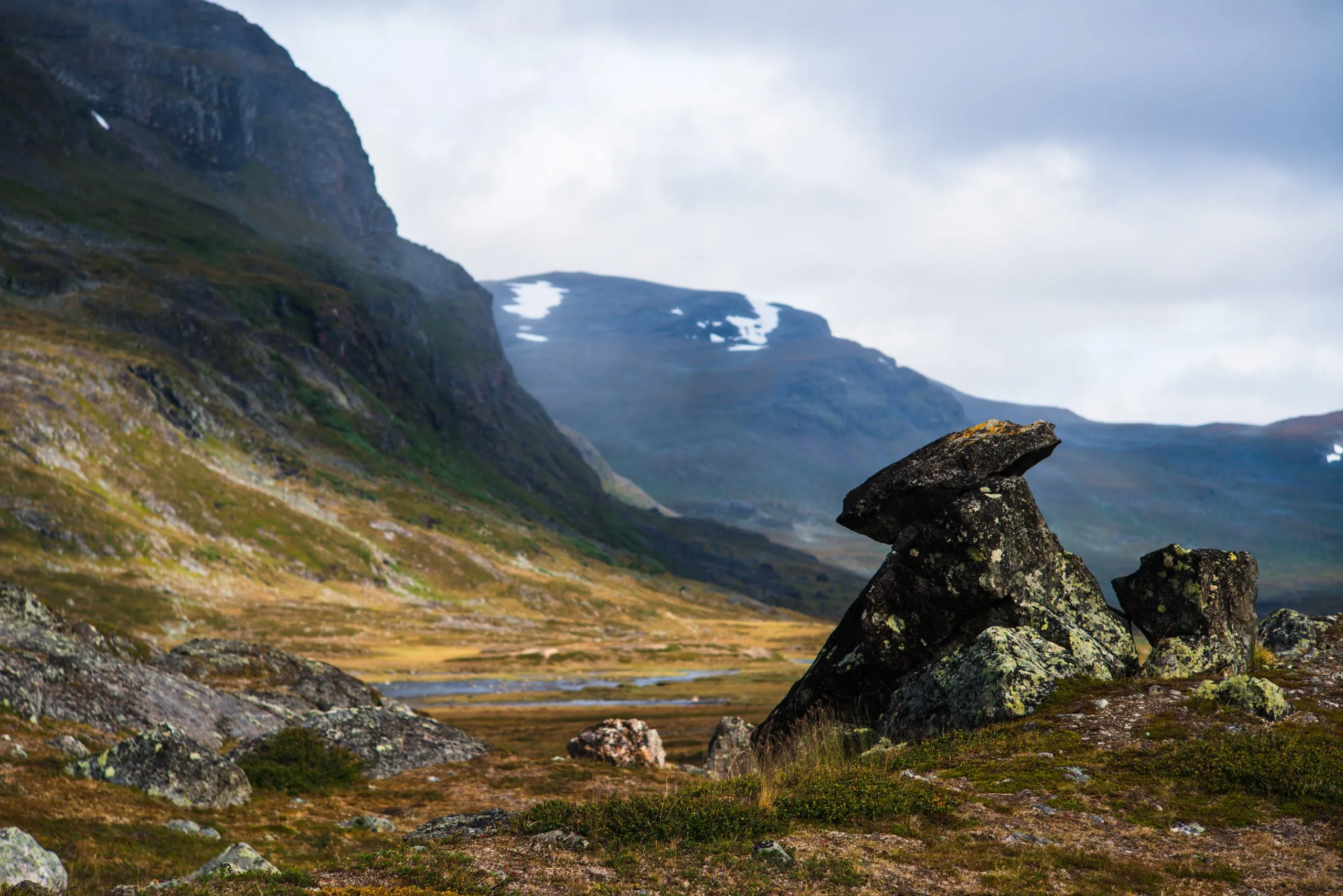 A pair of large lichen-covered glacial boulders sit on the moor of a Swedish Lapland valley, snow-streaked mountains rising behind.