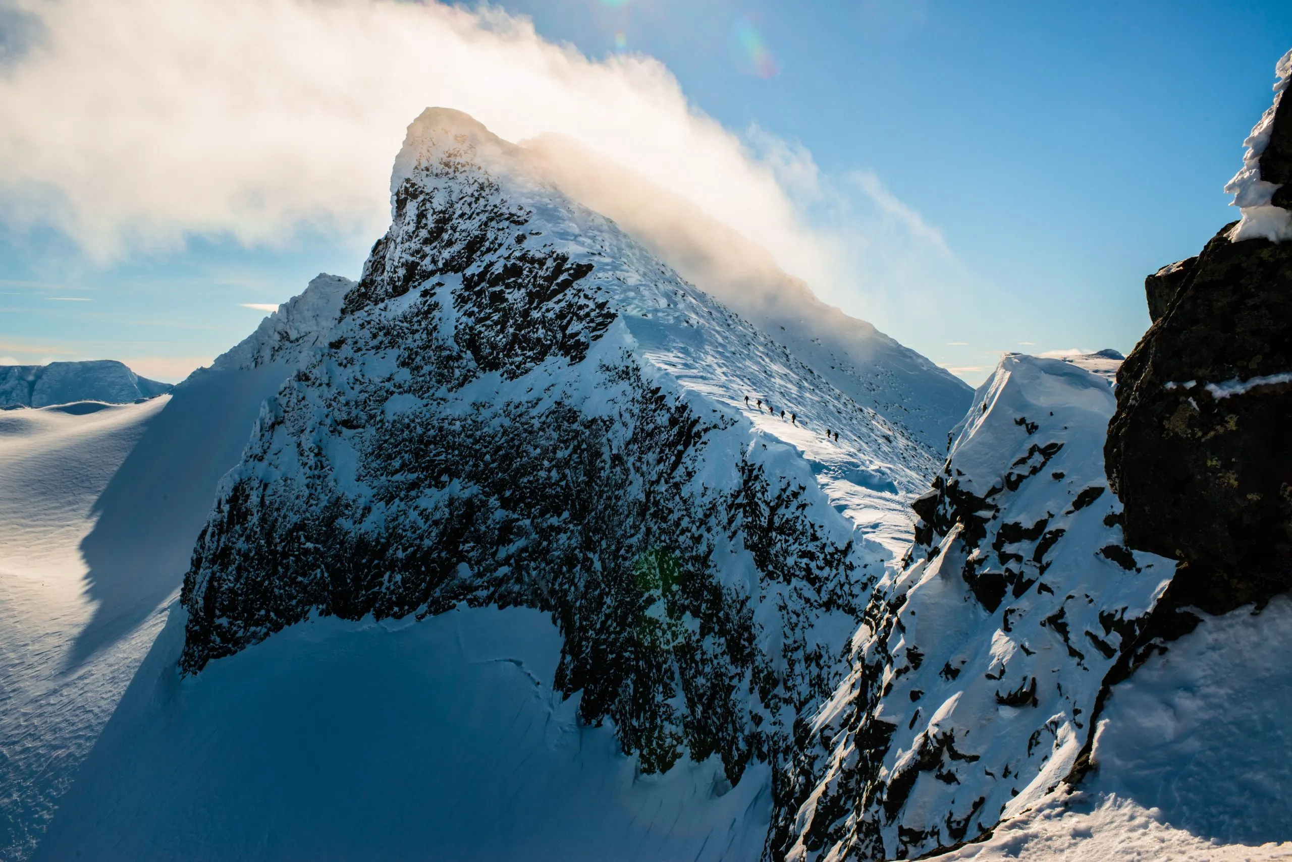 A wind-blown ridge of Kebnekaise in Swedish Lapland under cold winter sun, with a tiny line of climbers crossing the snowy crest.