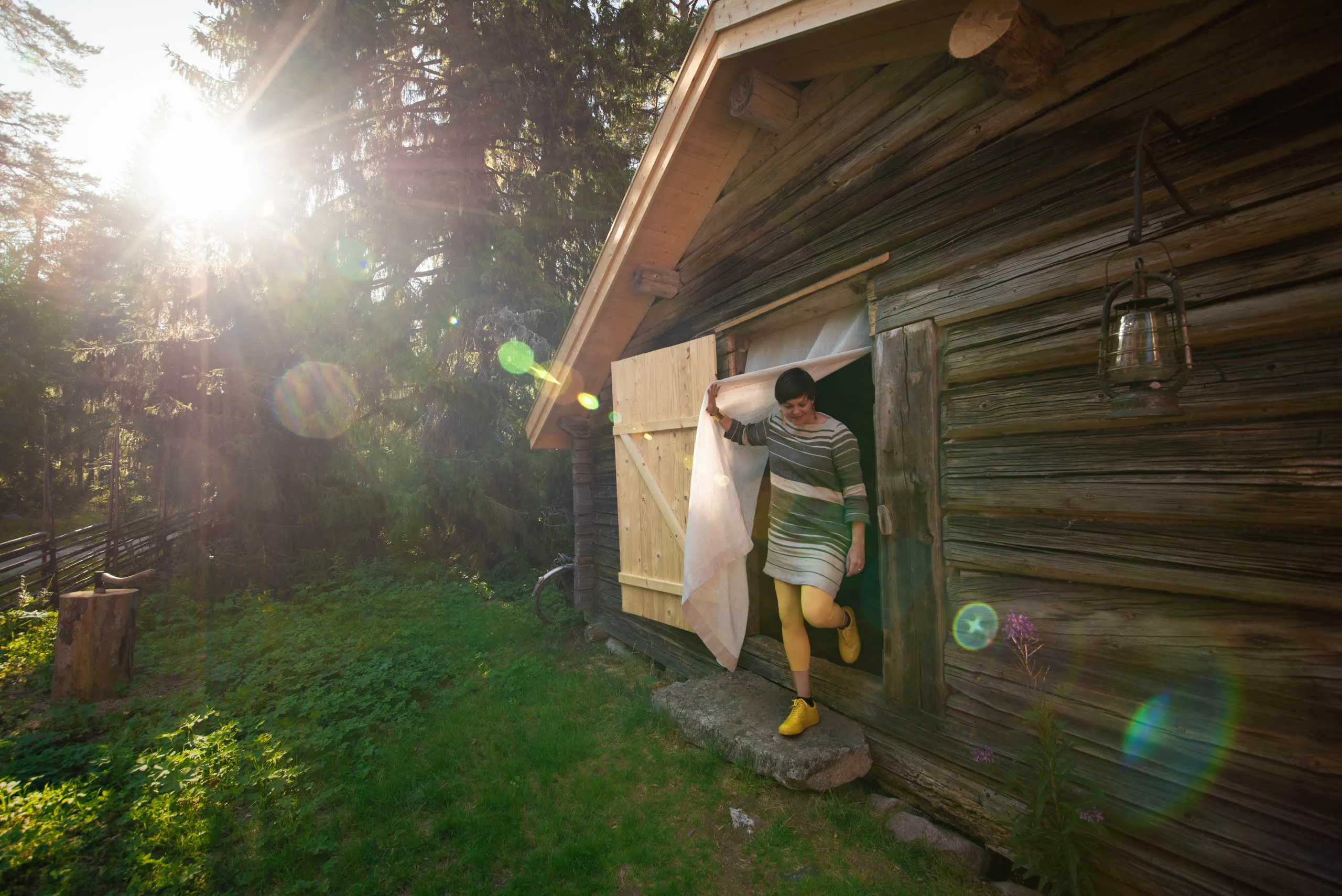 A woman in a striped dress and yellow tights steps out of a weathered timber cabin into golden forest light, holding a gauzy curtain aside.