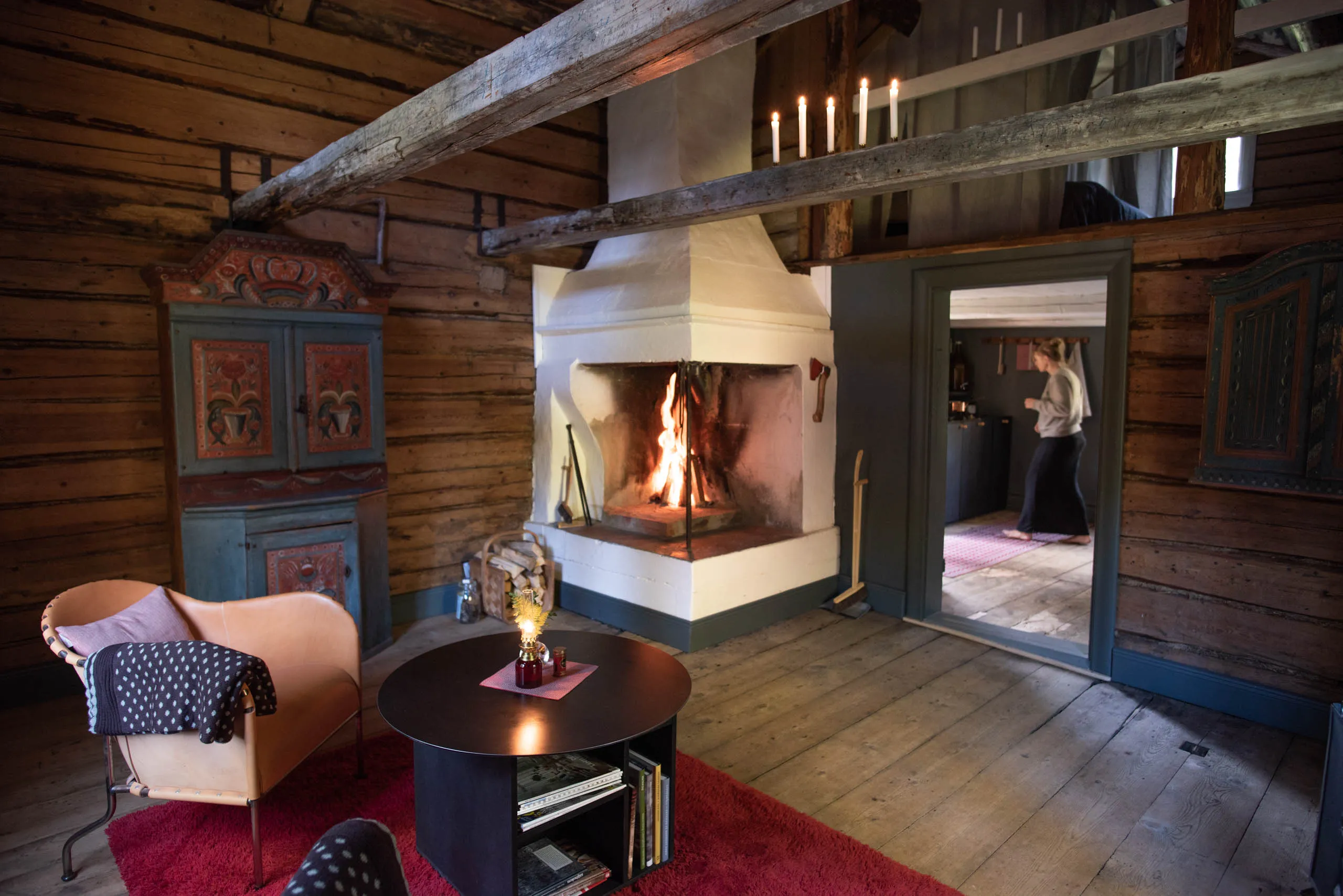 Cosy interior of a Hälsingland timber farmhouse with an open fire, a folk-painted kurbits cupboard and lit candles, a person visible through a doorway to the next room.