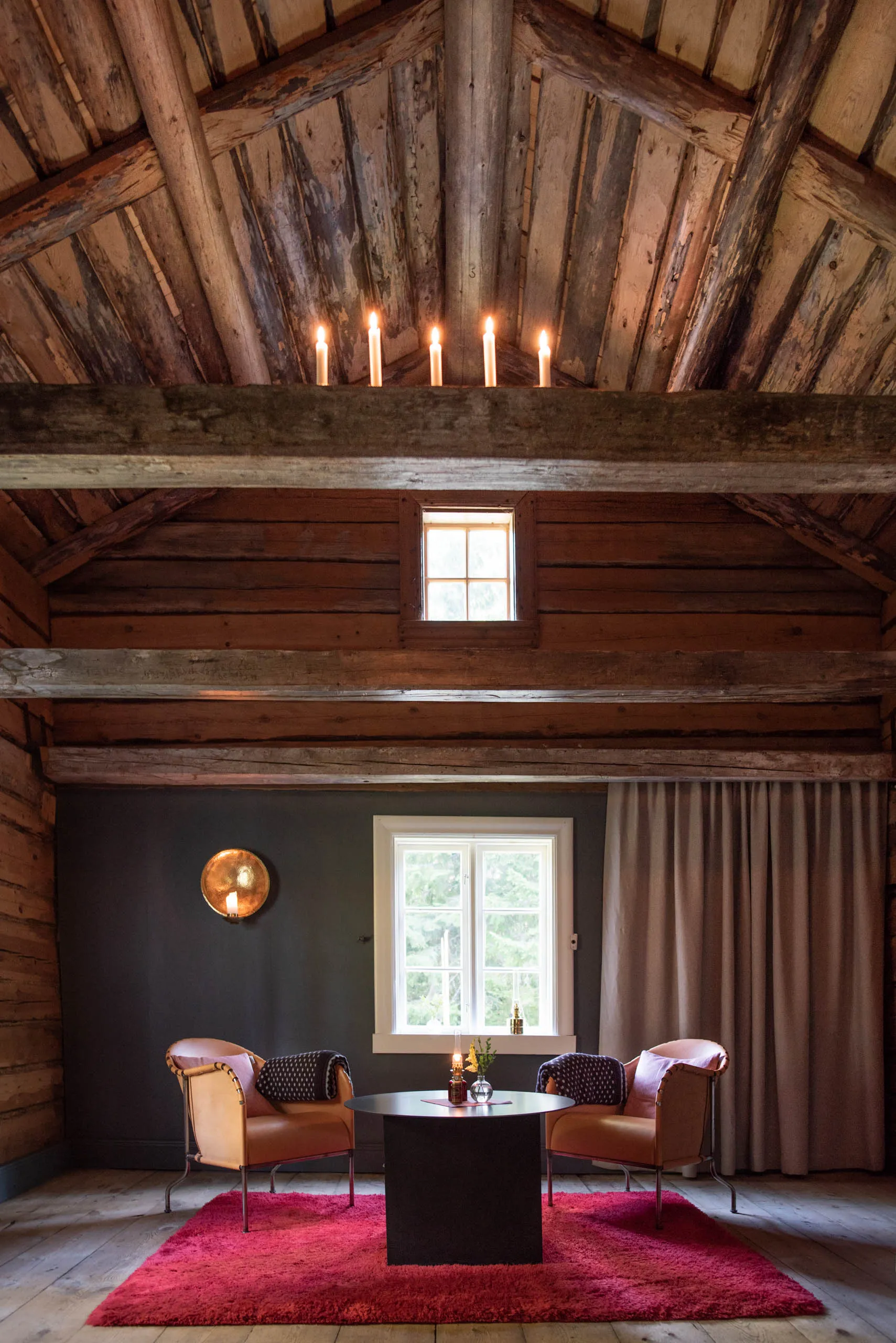 A vaulted timber loft room with exposed beams and lit candles overhead, two leather chairs and a small black table on a pink rug.
