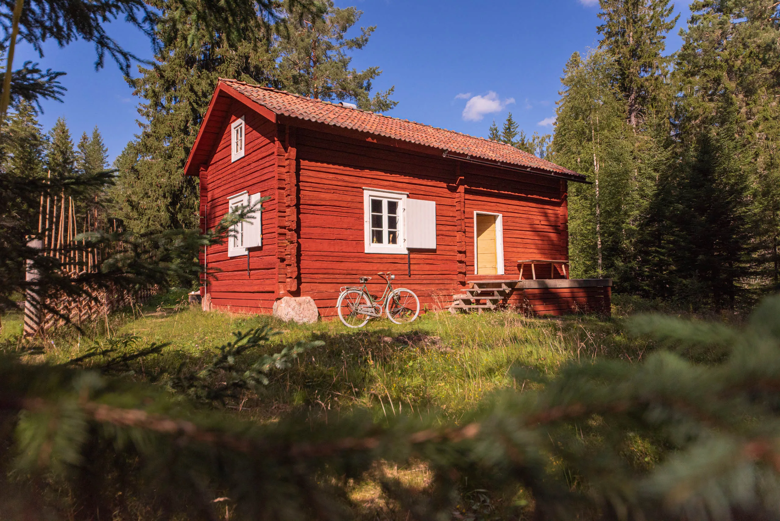 A classic falu-red Swedish timber cottage with white-trimmed windows in a forest clearing, a bicycle leaned against the wall under a clear blue sky.