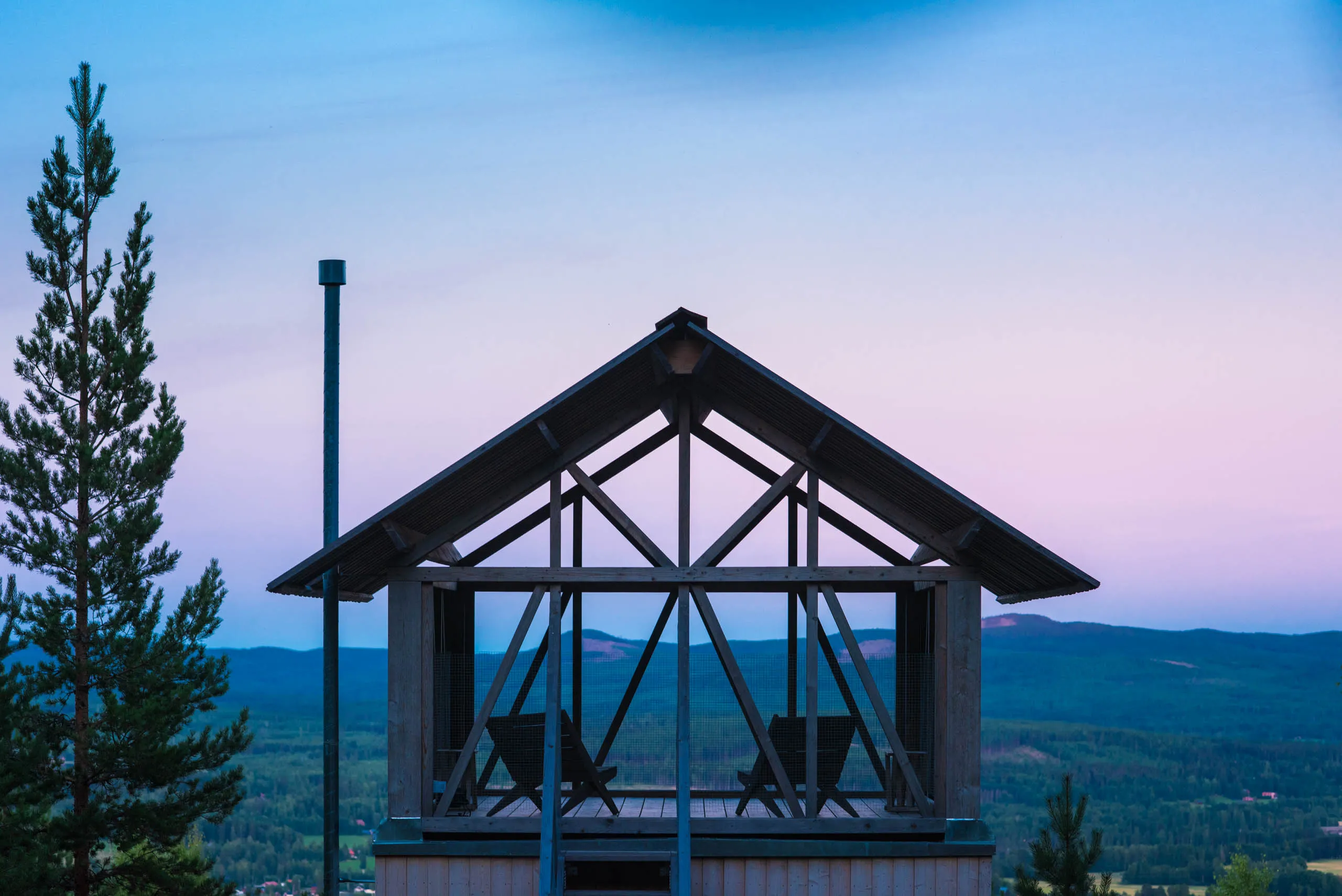 An open-frame wooden lookout shelter with two armchairs perched on a forested hilltop, the pink-and-blue twilight sky lighting the mountains behind.