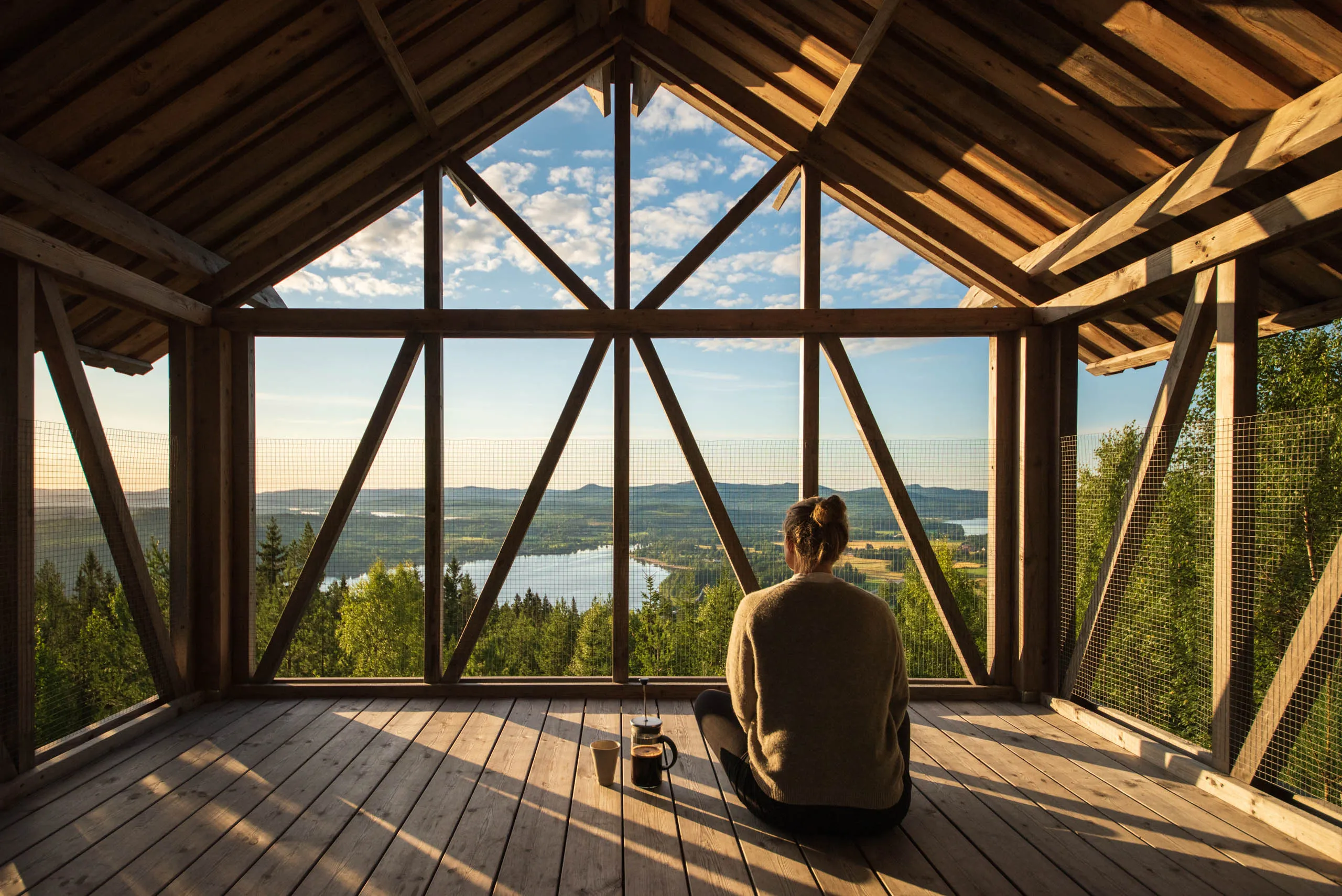 A woman sits on the floor of an open loft in the morning light, with freshly brewn coffee at her side, overlooking the mountains and forest of Orbaden in Hälsingland.