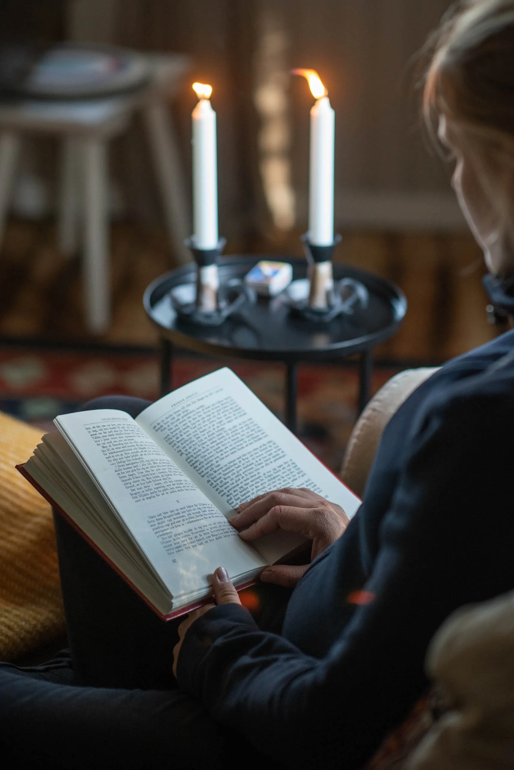A person reads a book beside two slim white candles on a black tray table in a softly lit Swedish living room.