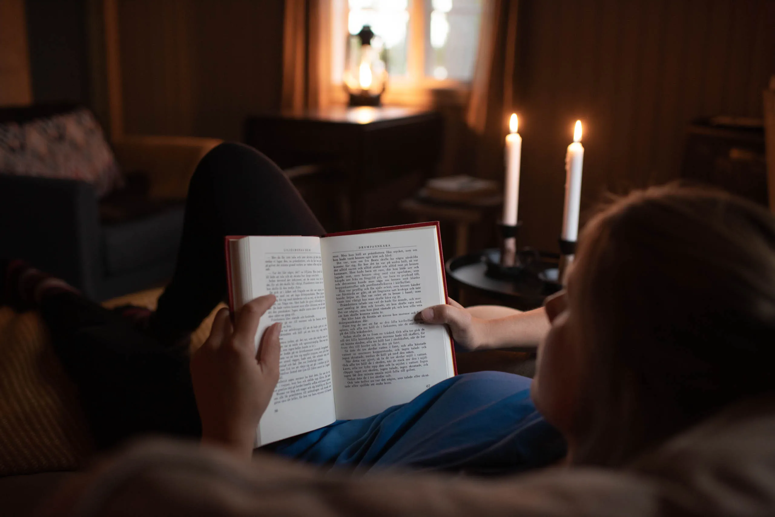 A reader curled up with a book in a candle-lit cabin room, a glowing oil lantern on the table behind and a window framed by curtains beyond.