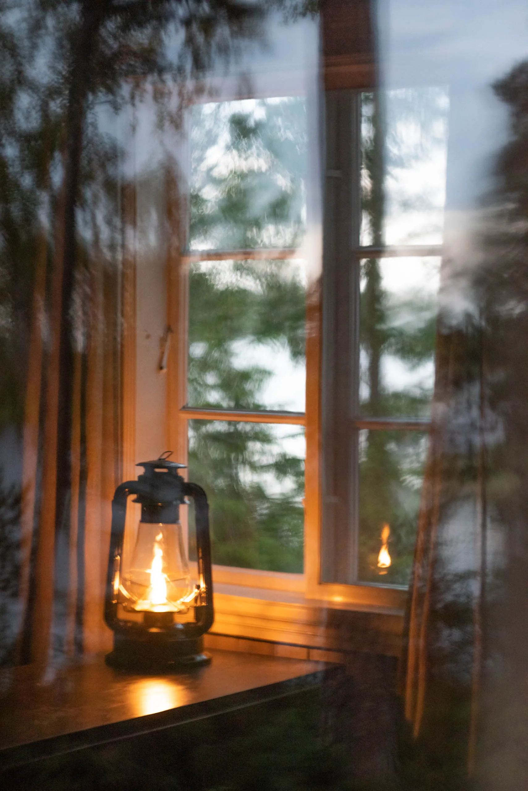 A lit oil lantern on a wooden window sill of a forest cabin at dusk, soft motion blur from breeze-stirred curtains.
