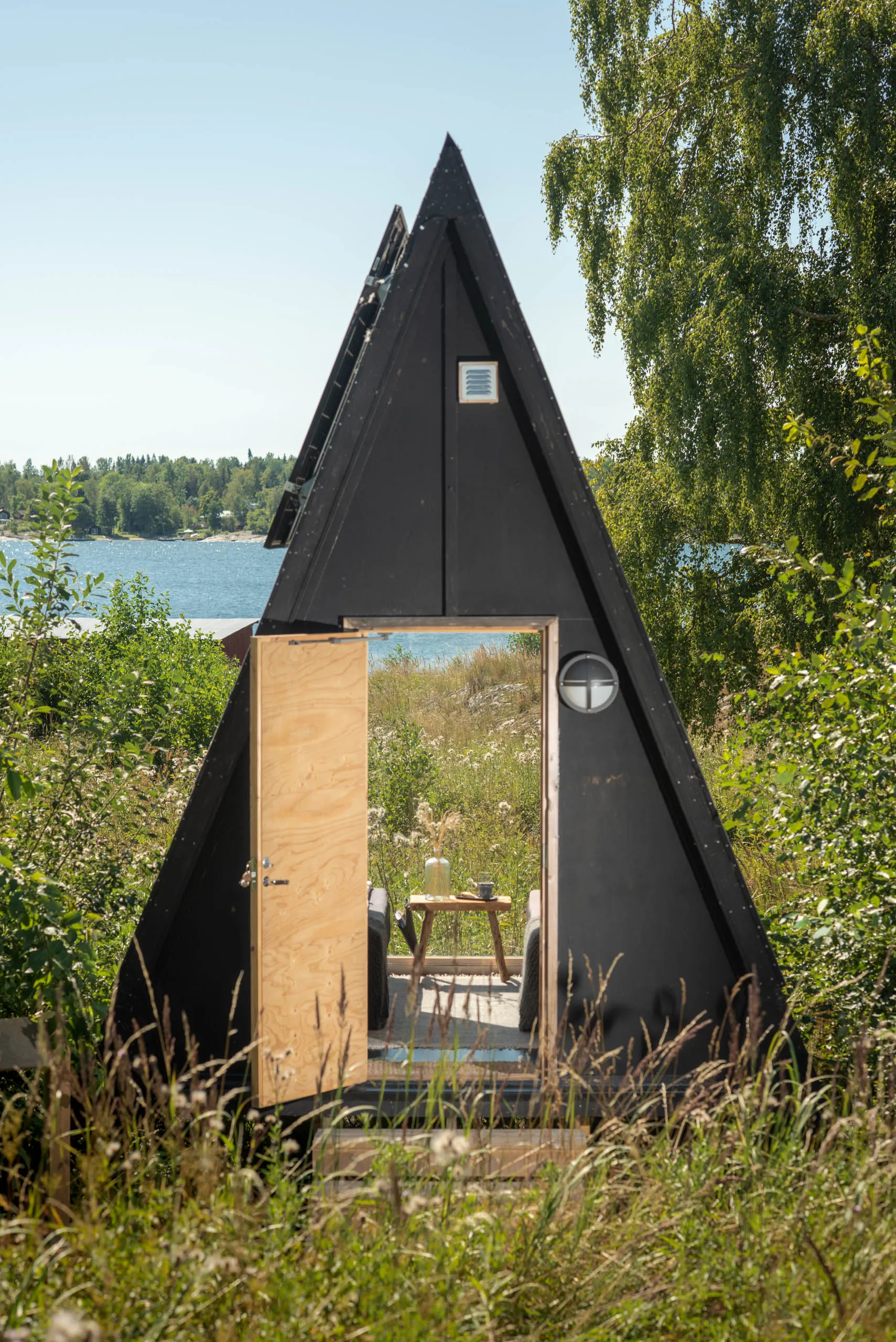 A small black wooden A-frame cabin stands in a lakeside meadow under a birch tree, its plywood door open to reveal a simple interior.