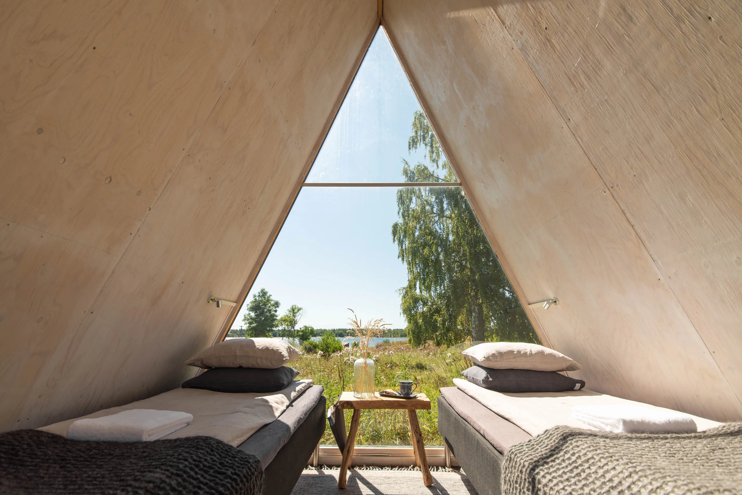Inside the same A-frame cabin: two single beds with linen pillows on either side of a tall triangular window framing the lake and a leaning birch.