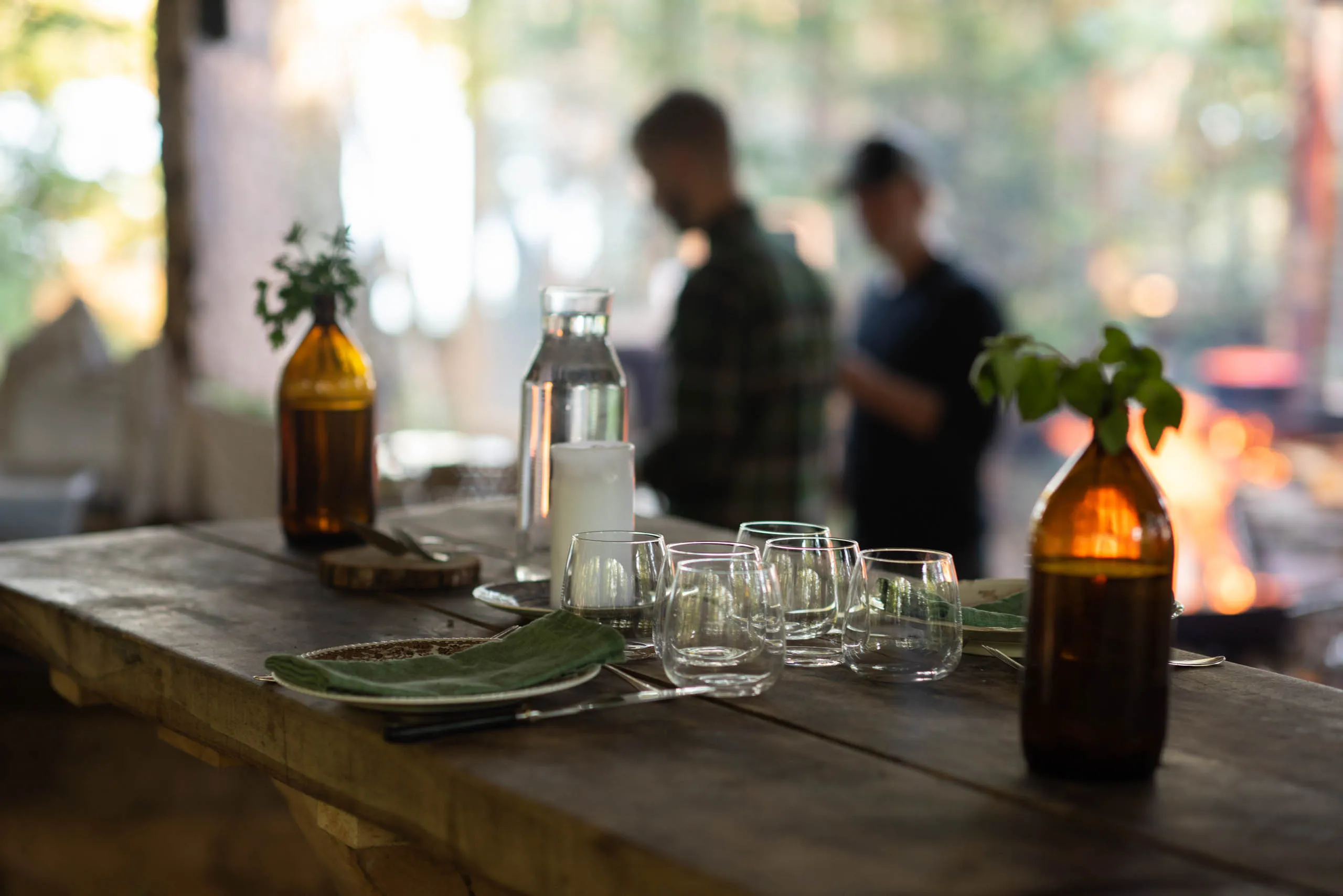 An outdoor table set with plates, glasses and bottles holding sprigs of greens, two figures preparing food beside a fire in the soft-focus background.