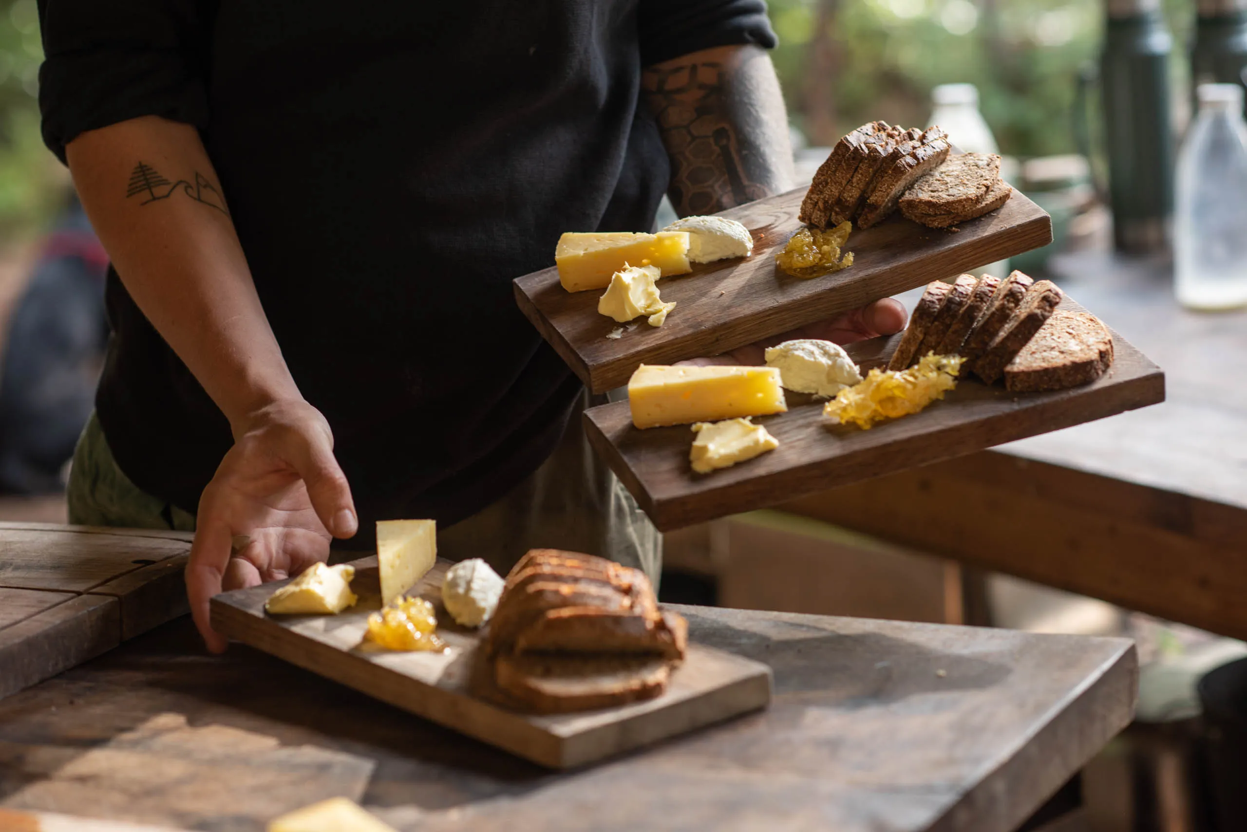 A tattooed forearm sets a wooden board of artisan cheeses, butter, honeycomb and dark rye bread onto a forest dining table.
