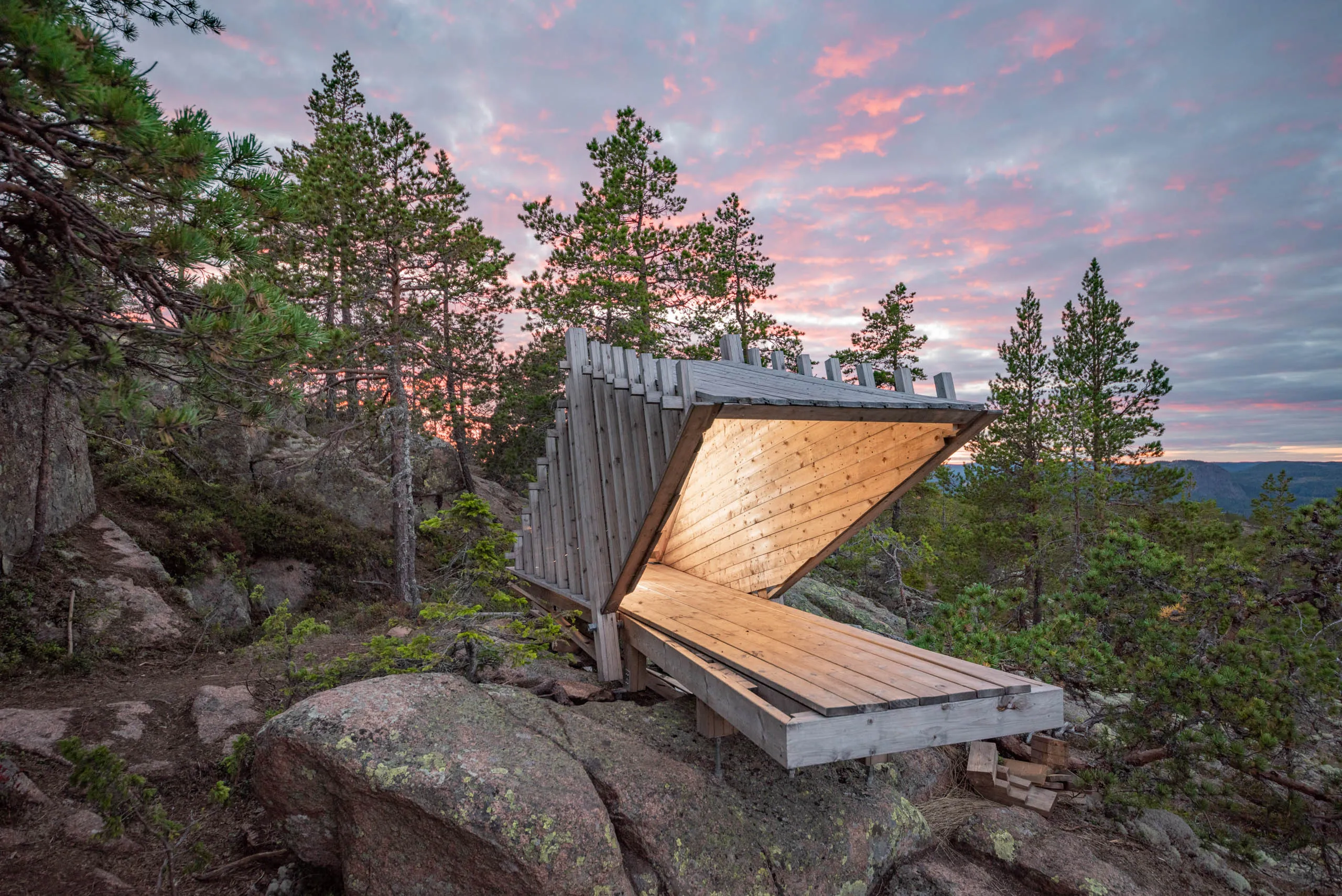 A hut or shack built with minimalistic architecture stands on top of a forested hill of the High Coast of Sweden, with a colorful twilight sky behind it.