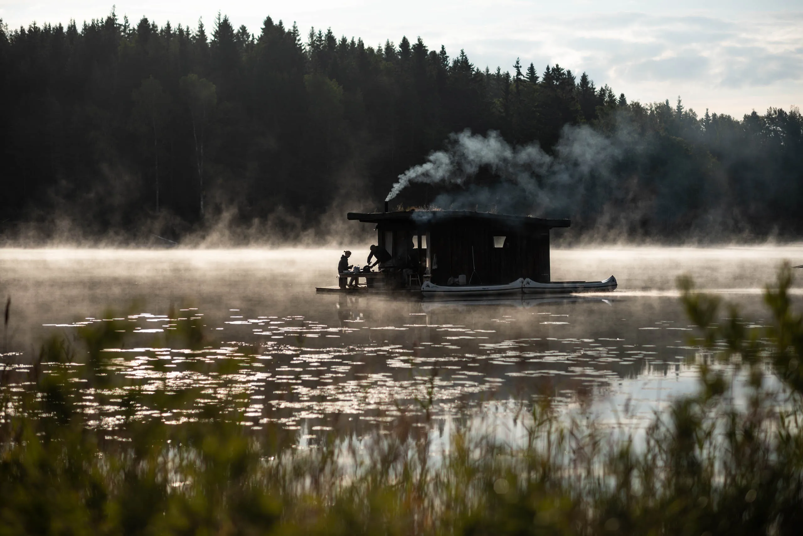 Two people on the deck of a floating sauna prepare a meal as smoke rises from the chimney, the lake mist lit gold by morning sun.