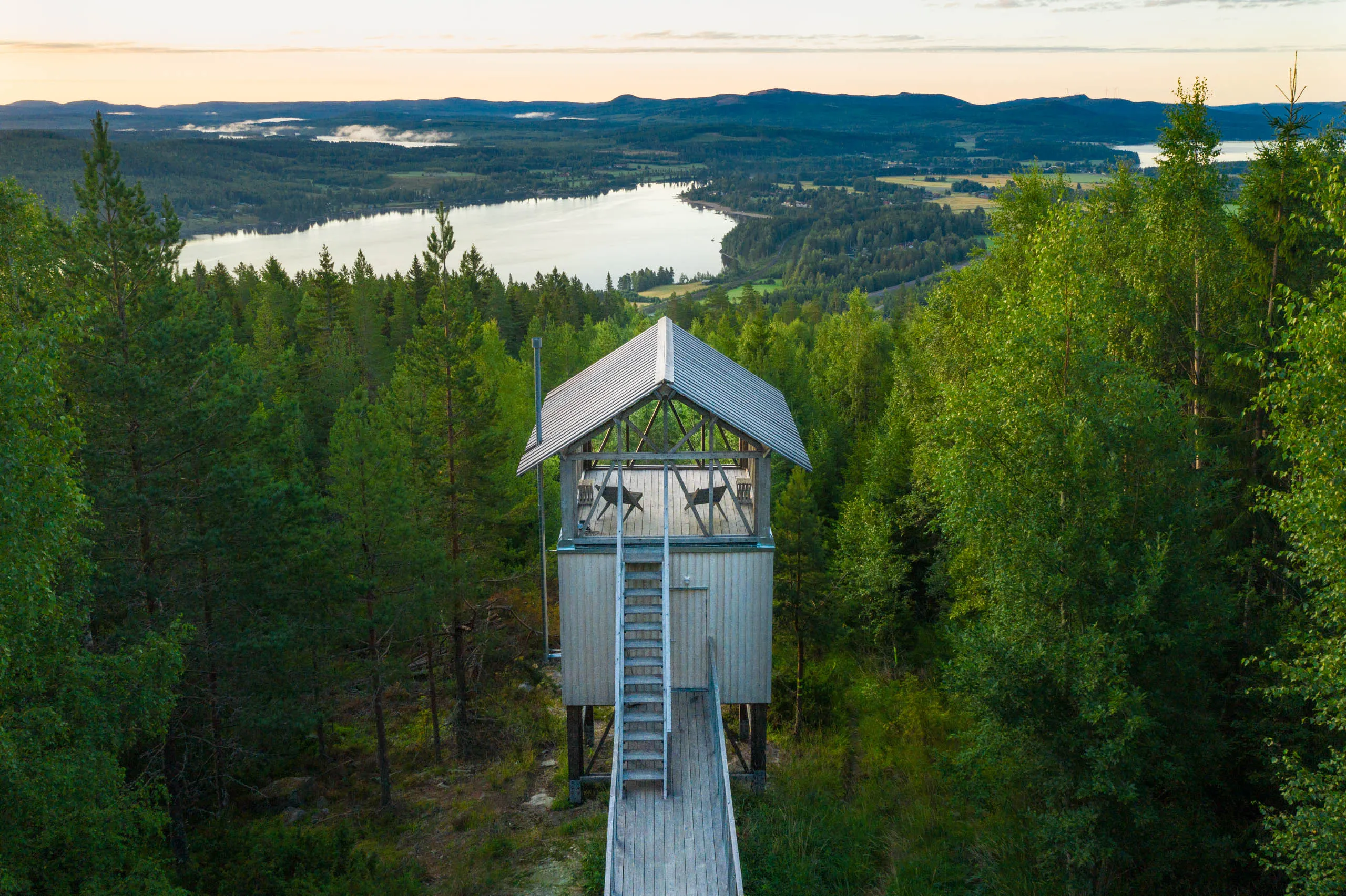 Drone view of a tall stilted wooden lookout cabin on a forested hilltop with a long boardwalk approach, the lake and farmland of Hälsingland behind.