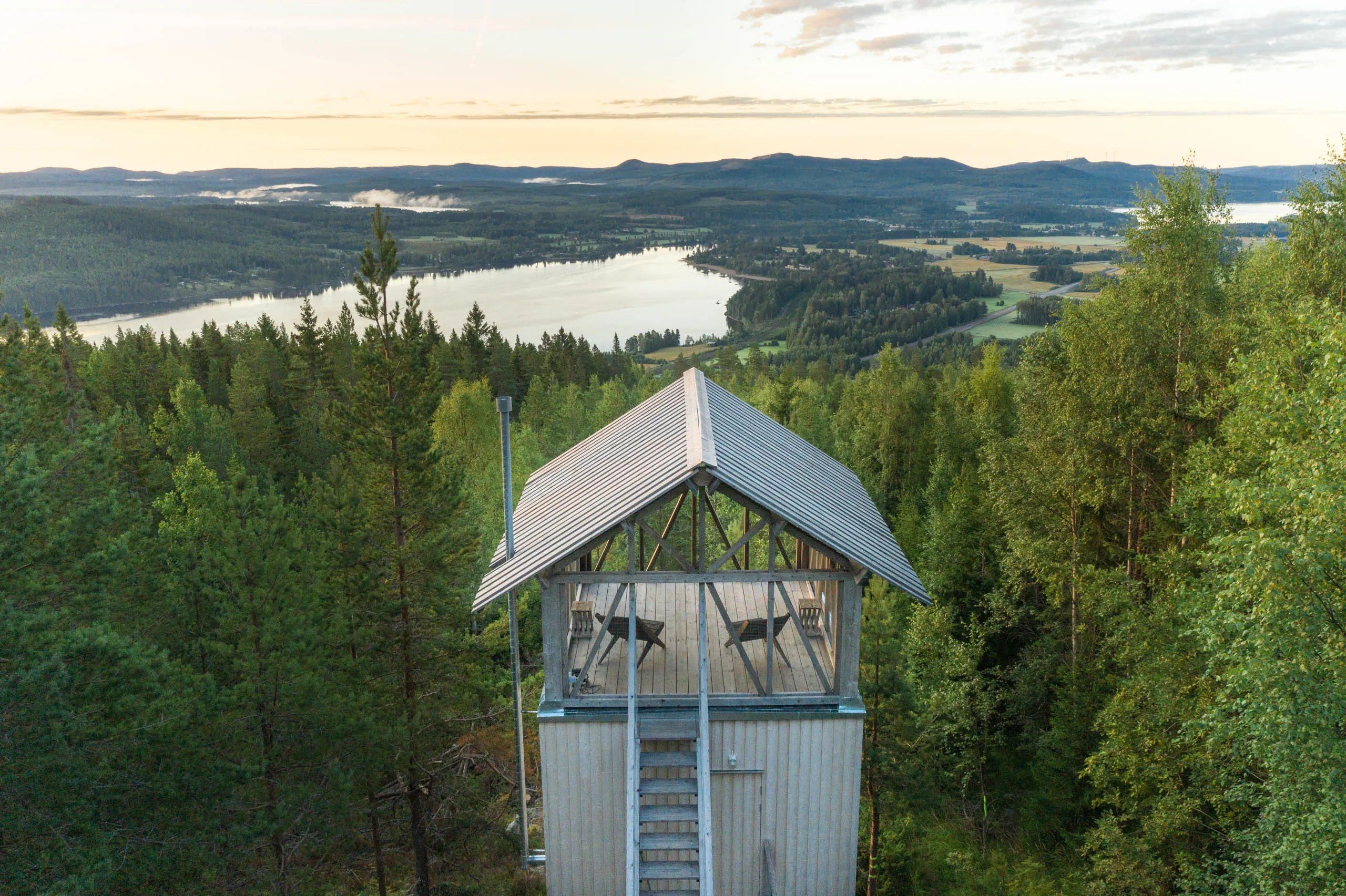 Drone view of the same hilltop lookout cabin from a closer angle, dense pines around it and a long lake stretching toward the horizon.