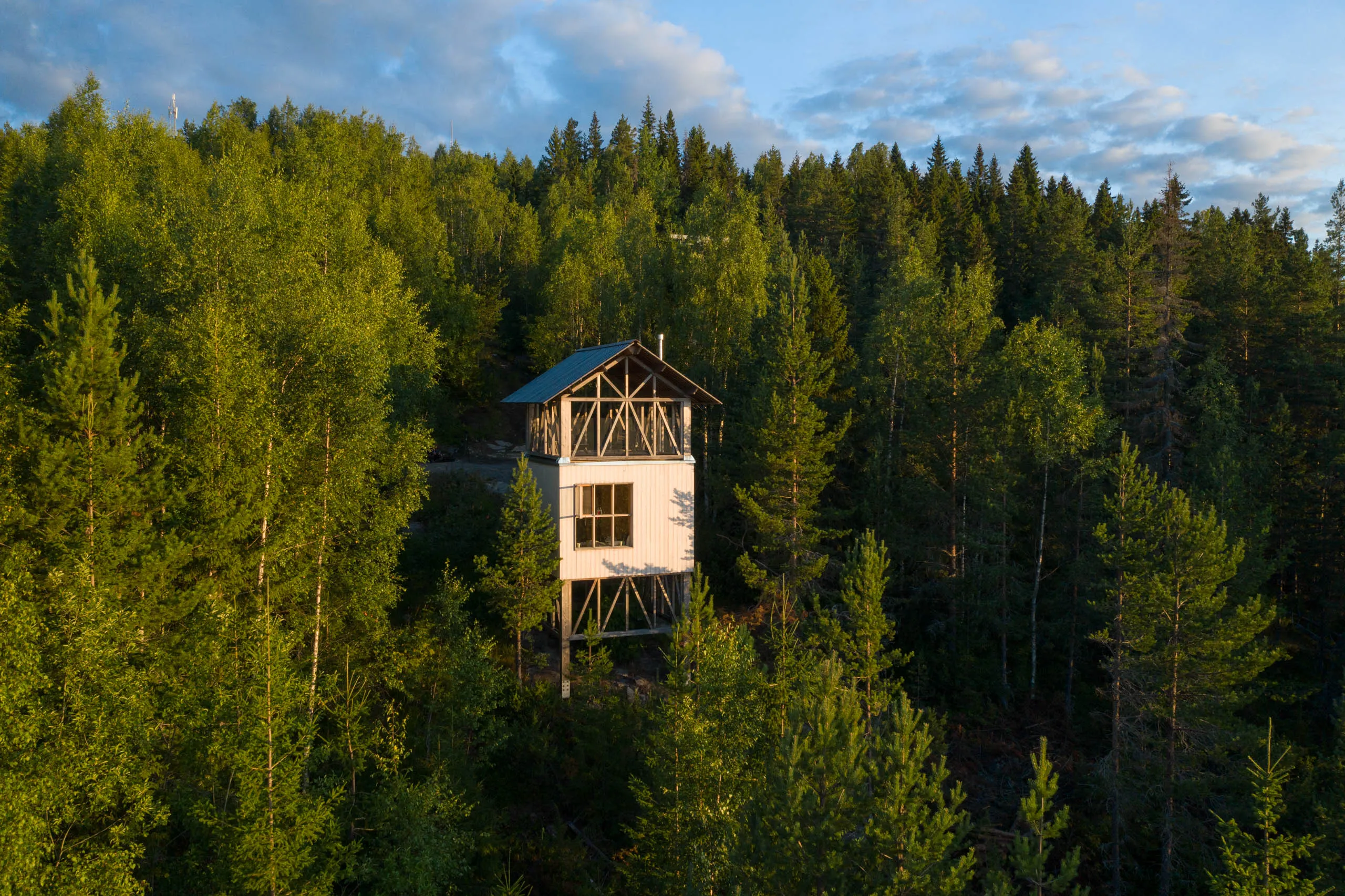 An elevated three-storey wooden cabin perched on stilts in a Swedish pine forest, viewed from above by drone.