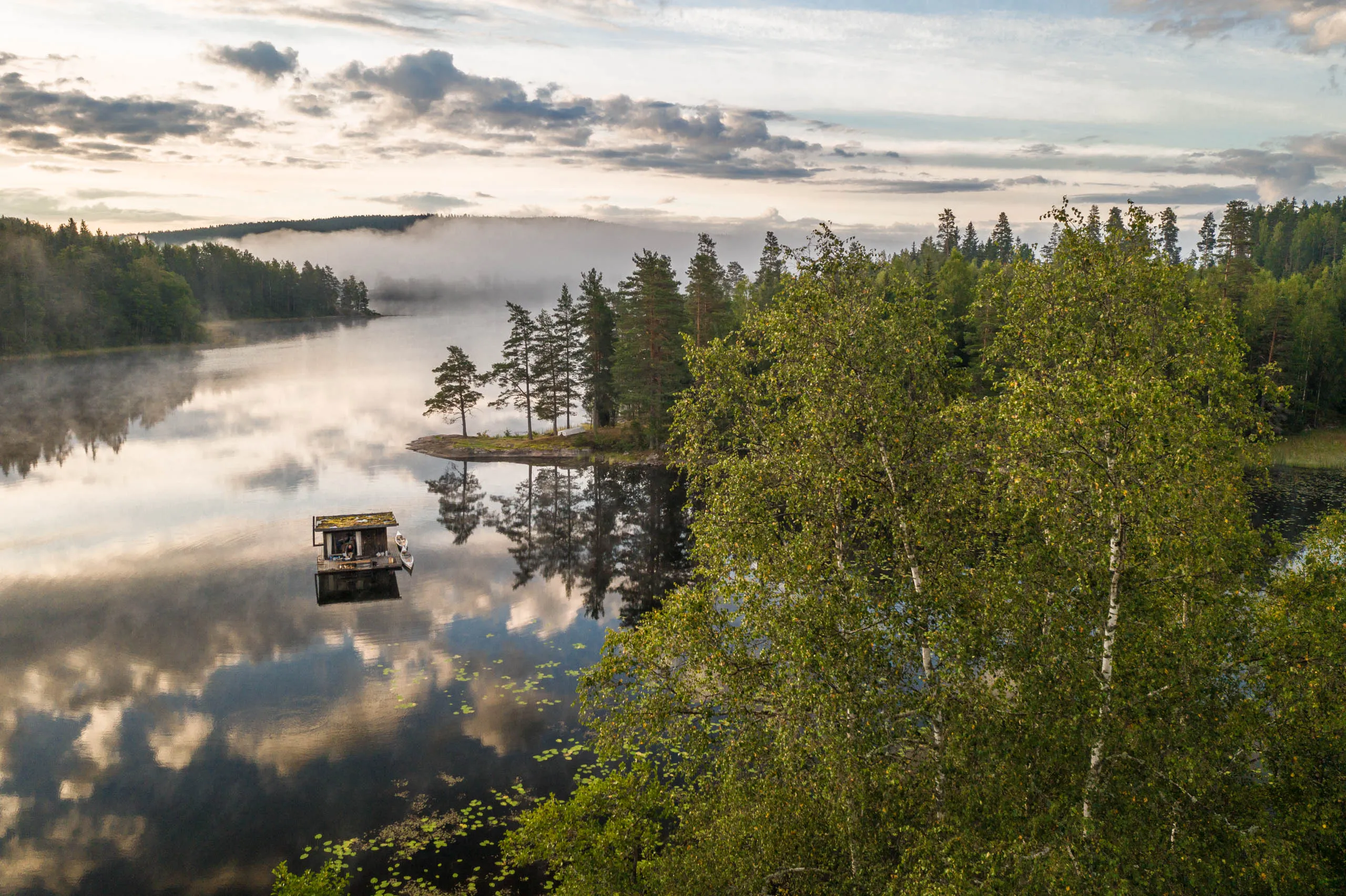 Drone view of the floating sauna on a misty Swedish lake at dawn, the wooden cabin reflected in the still water with birches in the foreground.