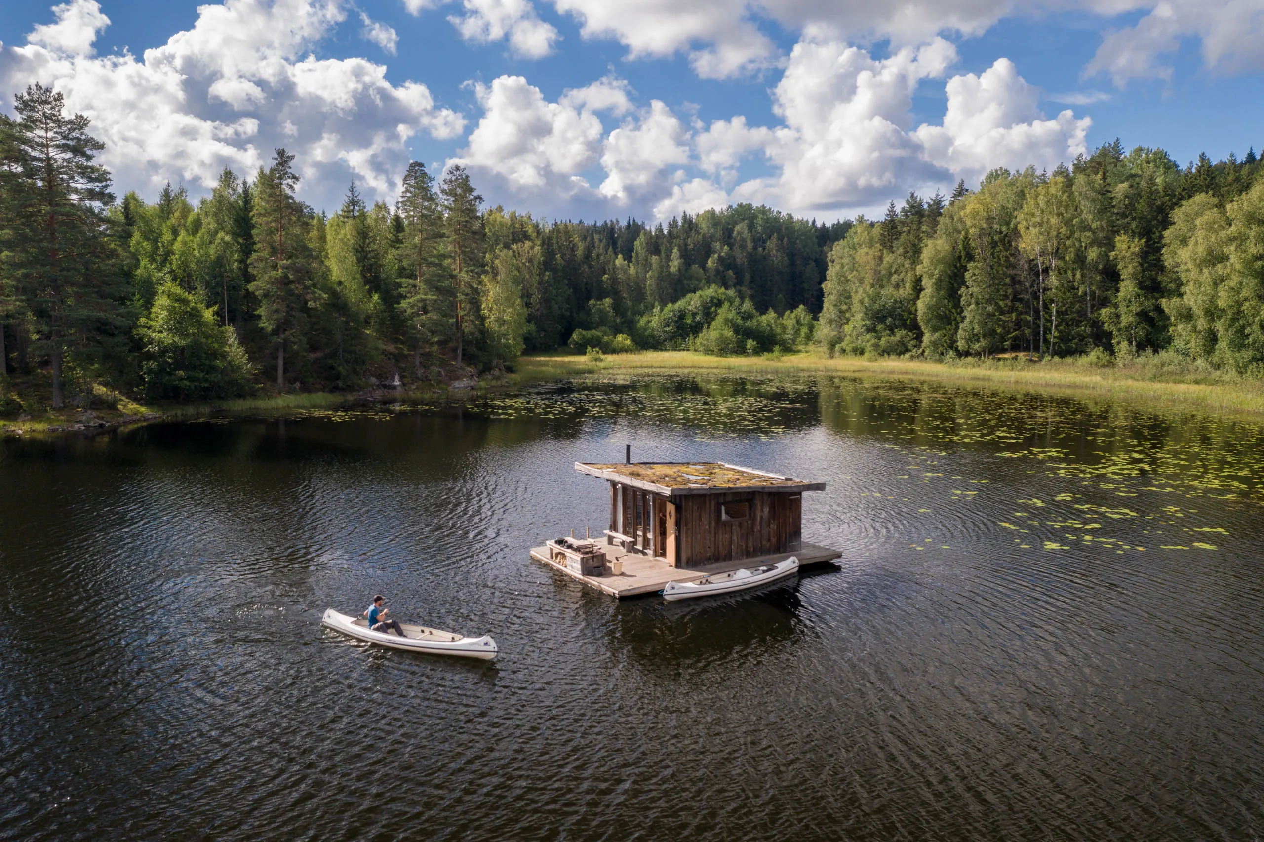 Aerial view of a floating sauna in the middle of a forest-rimmed lake, a person paddling a canoe up alongside in midday sun.