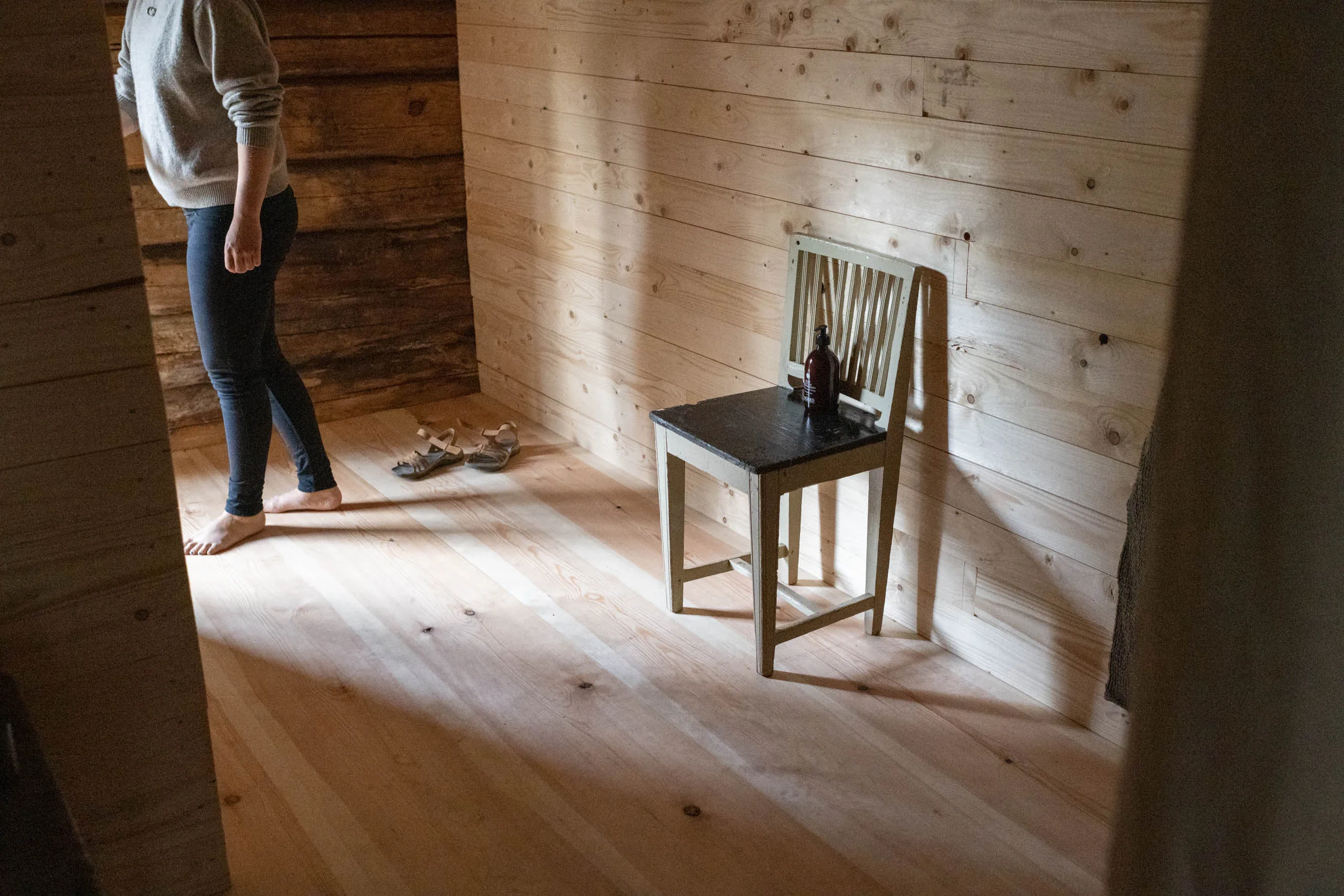 A barefoot person in jeans and a grey sweater stands in a sun-lit timber room with sandals on the floor and a small painted wooden chair holding a soap bottle.