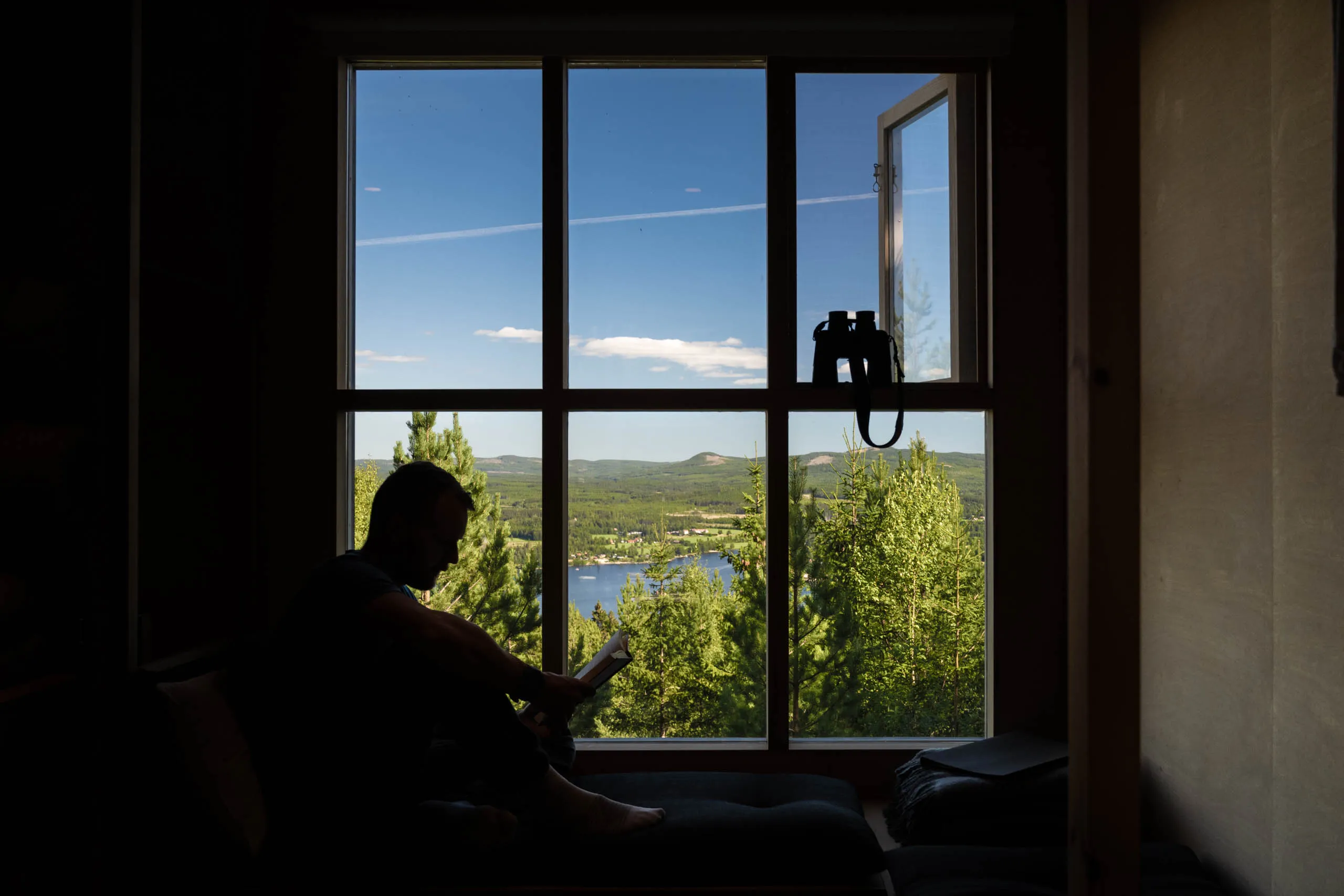 Silhouette of a person reading by a tall multi-pane window with a view over forested hills and a blue Swedish lake, binoculars hung on the open sash.