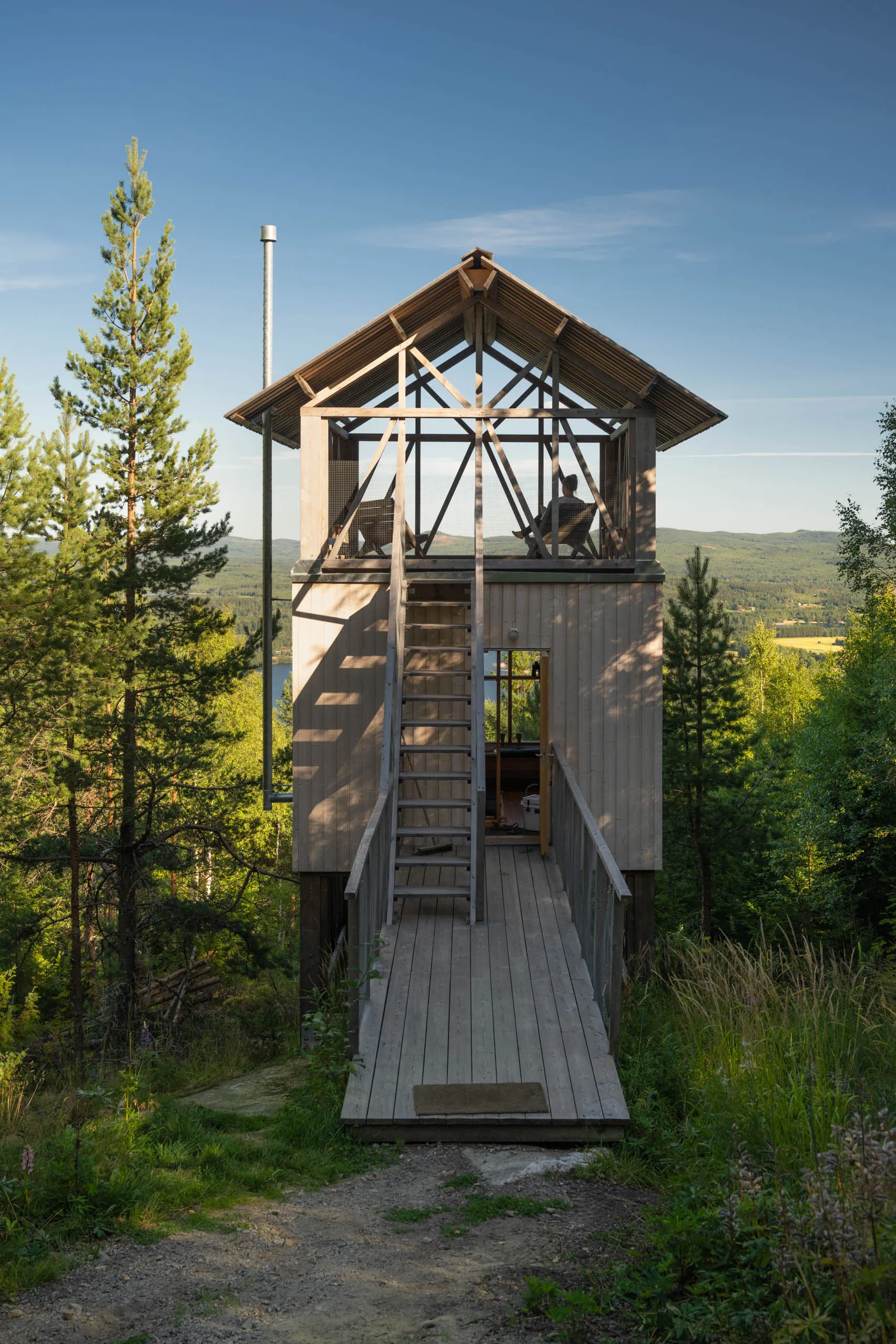 Tall slim wooden lookout cabin with an open-frame upper terrace and a long boardwalk approach, set on a forested hillside.