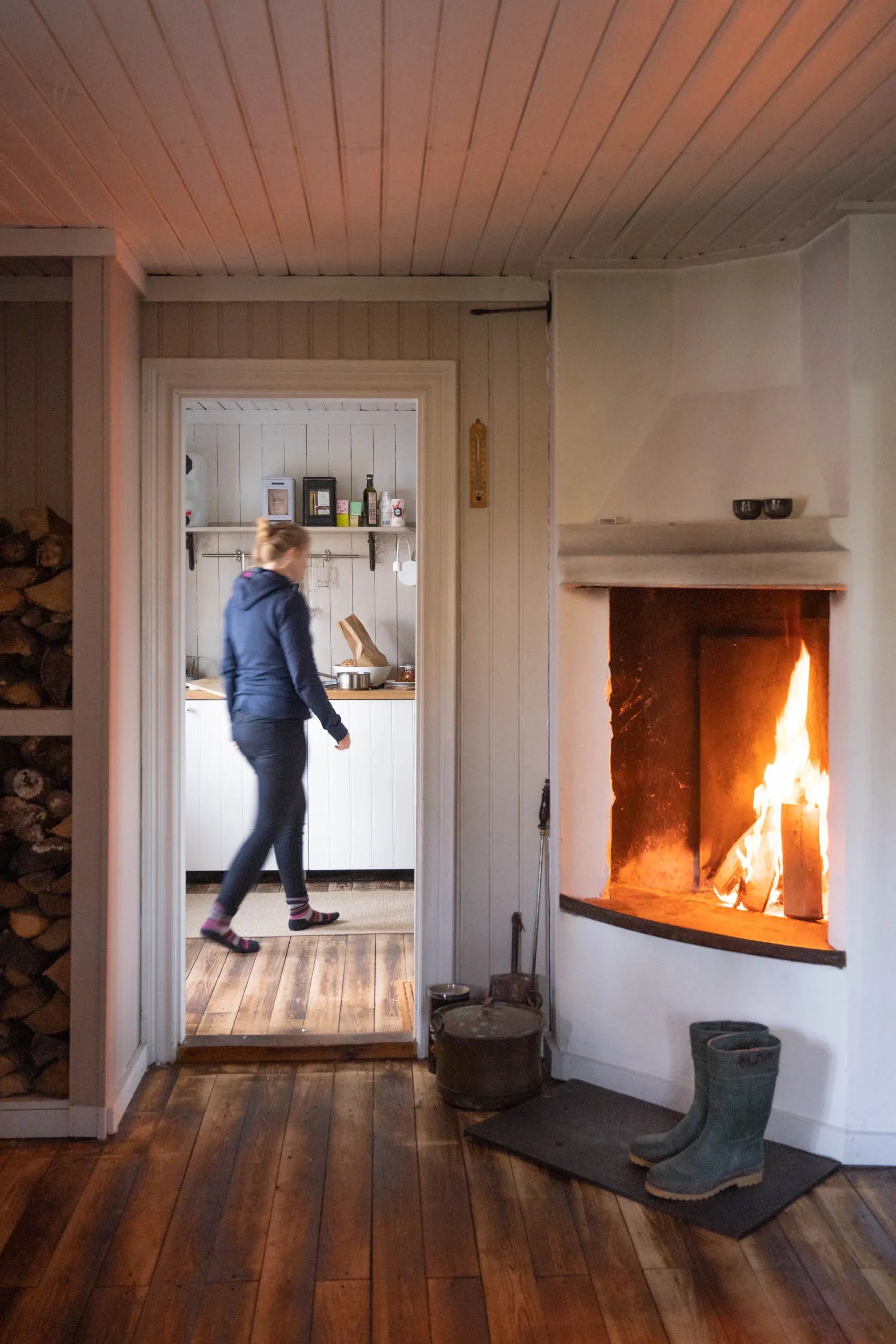 Cosy cabin interior with stacked firewood, a glowing tile-stove fireplace and blue rubber boots on the hearth, a woman walking through the doorway to a small kitchen beyond.