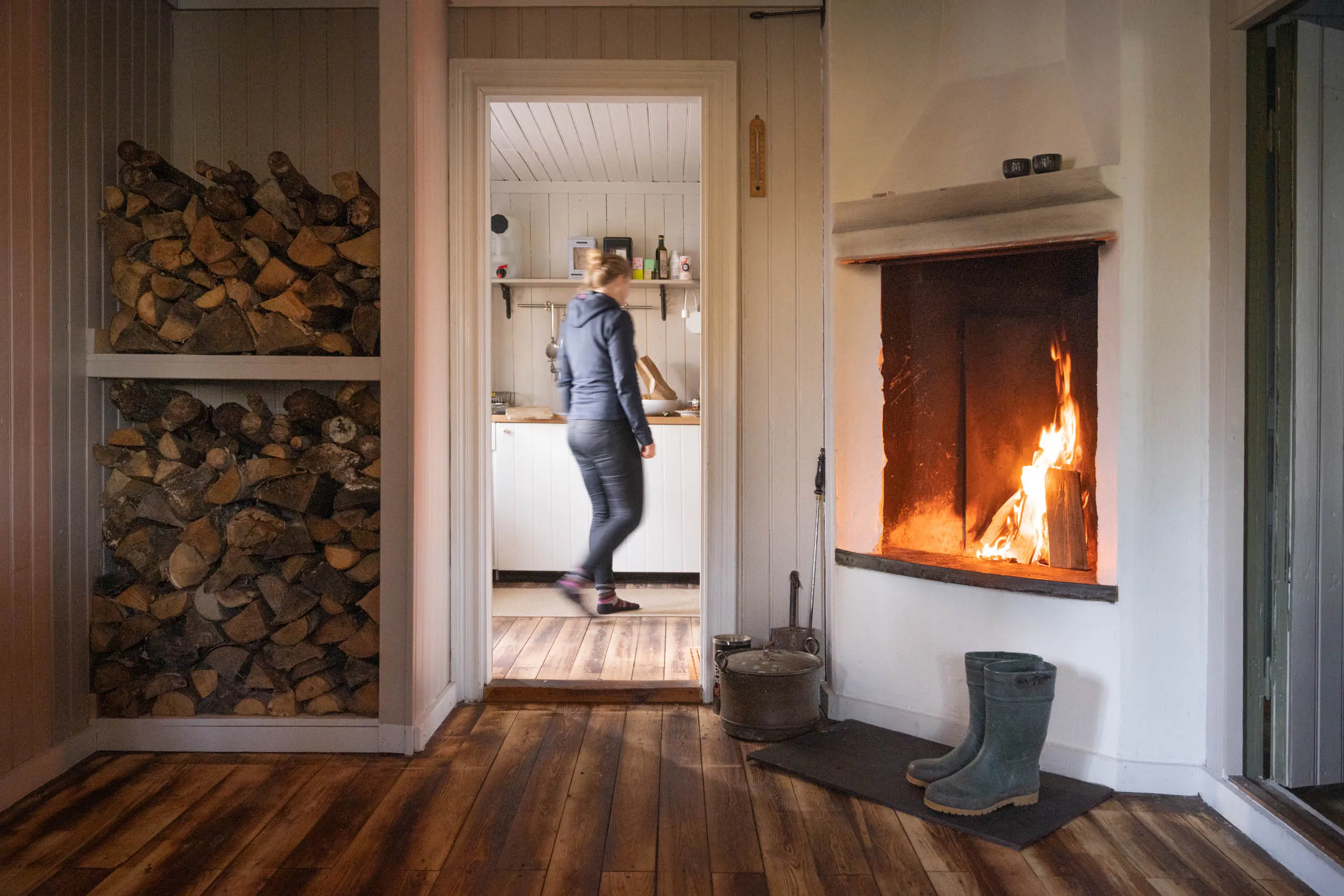 The same cabin scene from a wider angle, the firewood stack and tile-stove fire in the foreground and the kitchen beyond the doorway.
