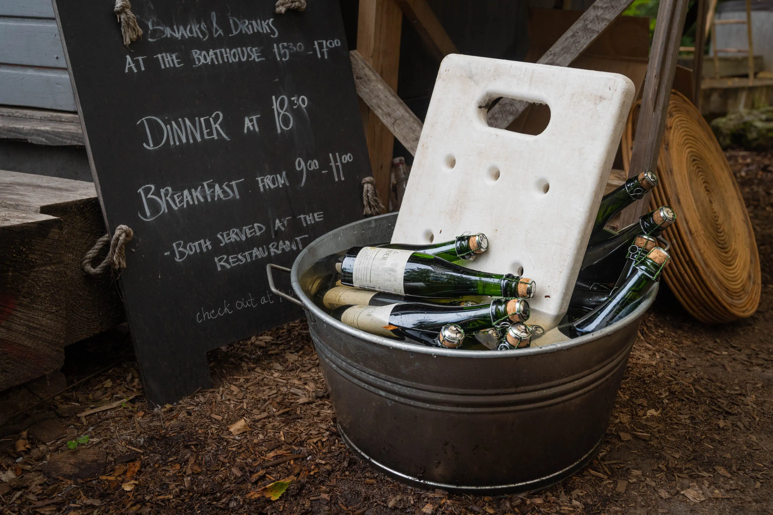A galvanised tub of wine and sparkling-drink bottles in front of a chalkboard listing meal times for a forest restaurant.