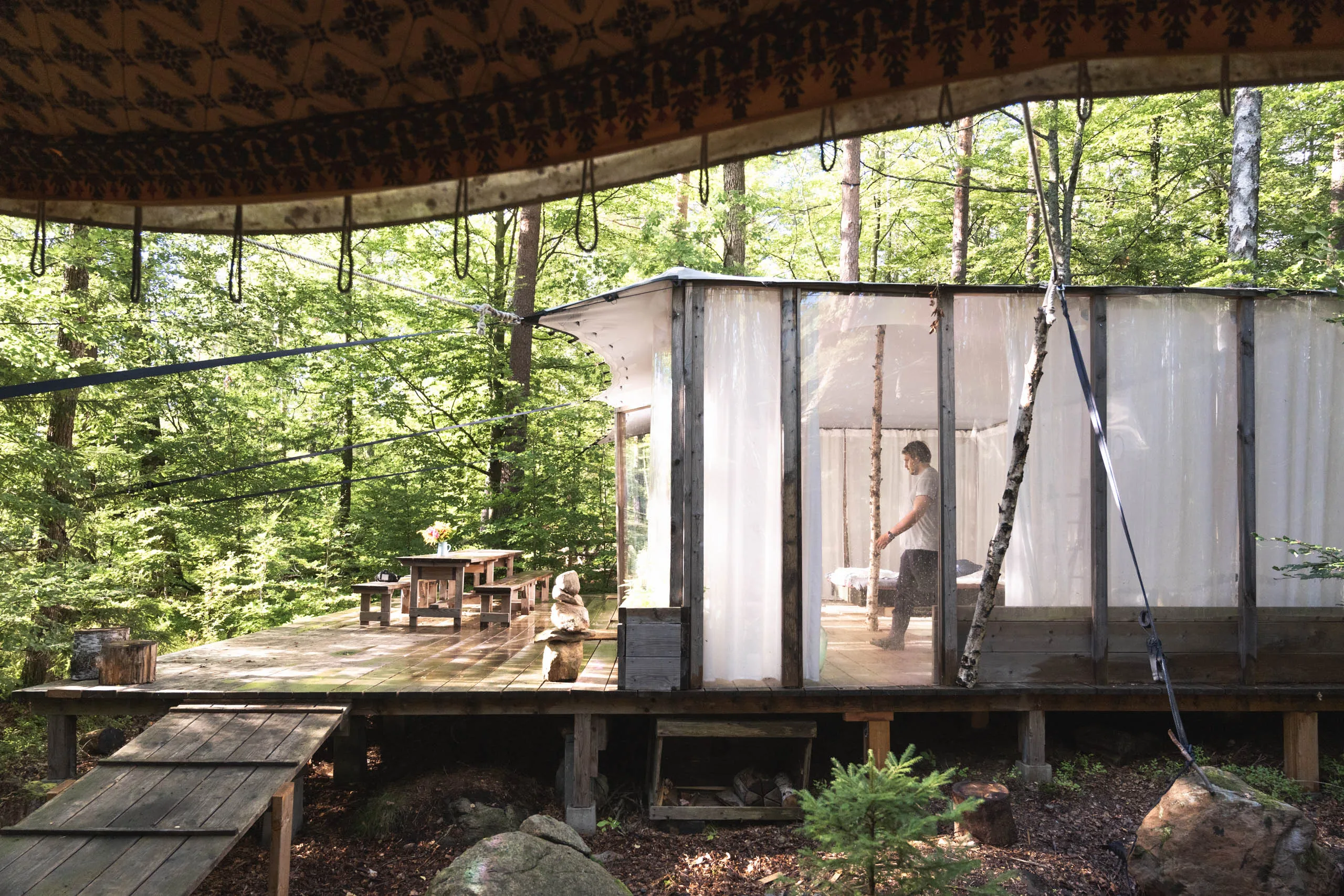 An open-air dining shelter with translucent plastic curtain walls in a beech forest, a person inside setting up beside the long table.